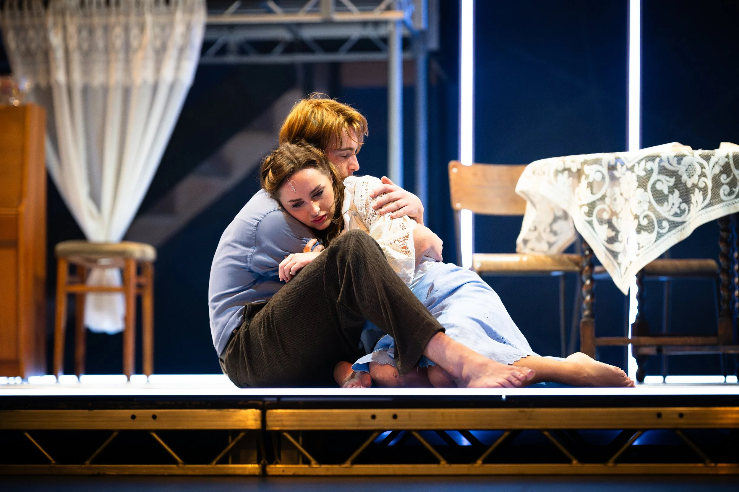 Two actors sitting on a stage, with one actor embracing the other in a scene from a play.