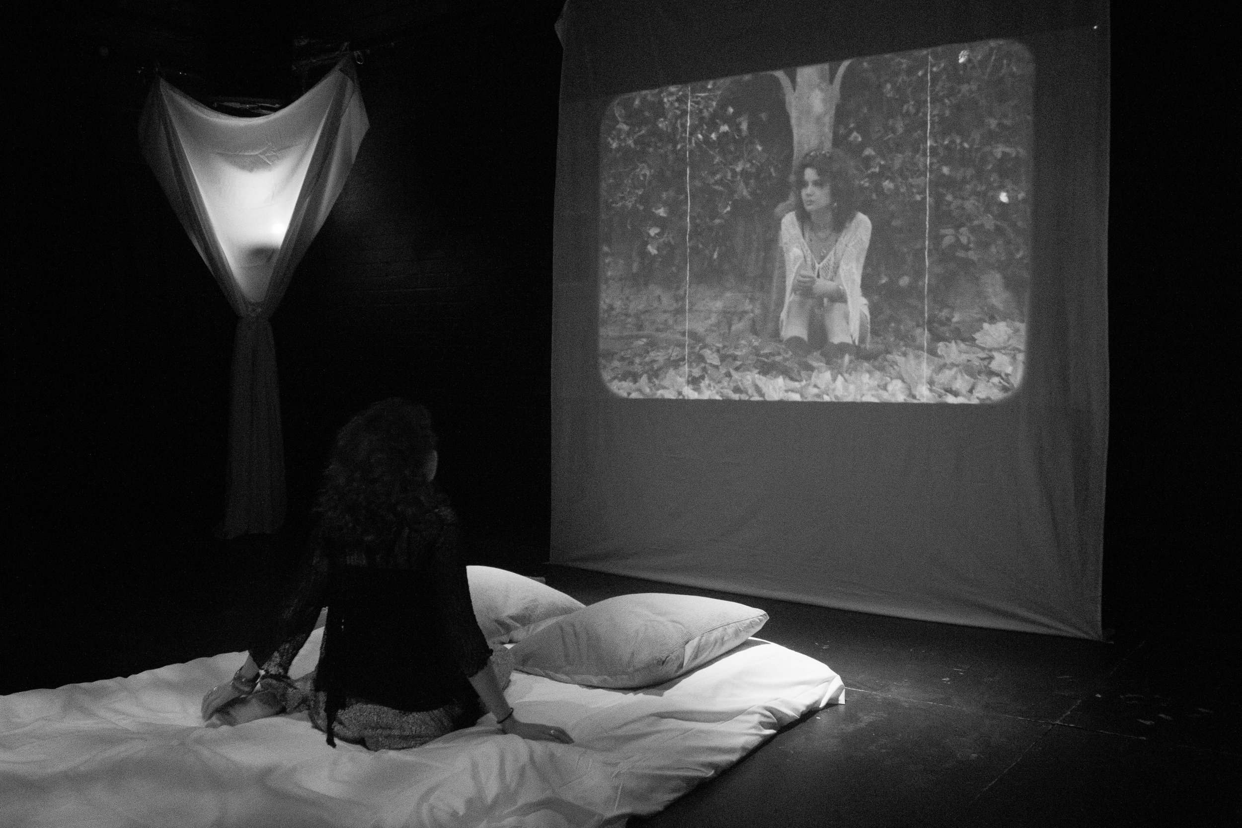 A person sitting cross-legged on a white cushion, watching a black-and-white projected image of a woman sitting outdoors with trees and leaves in the background.