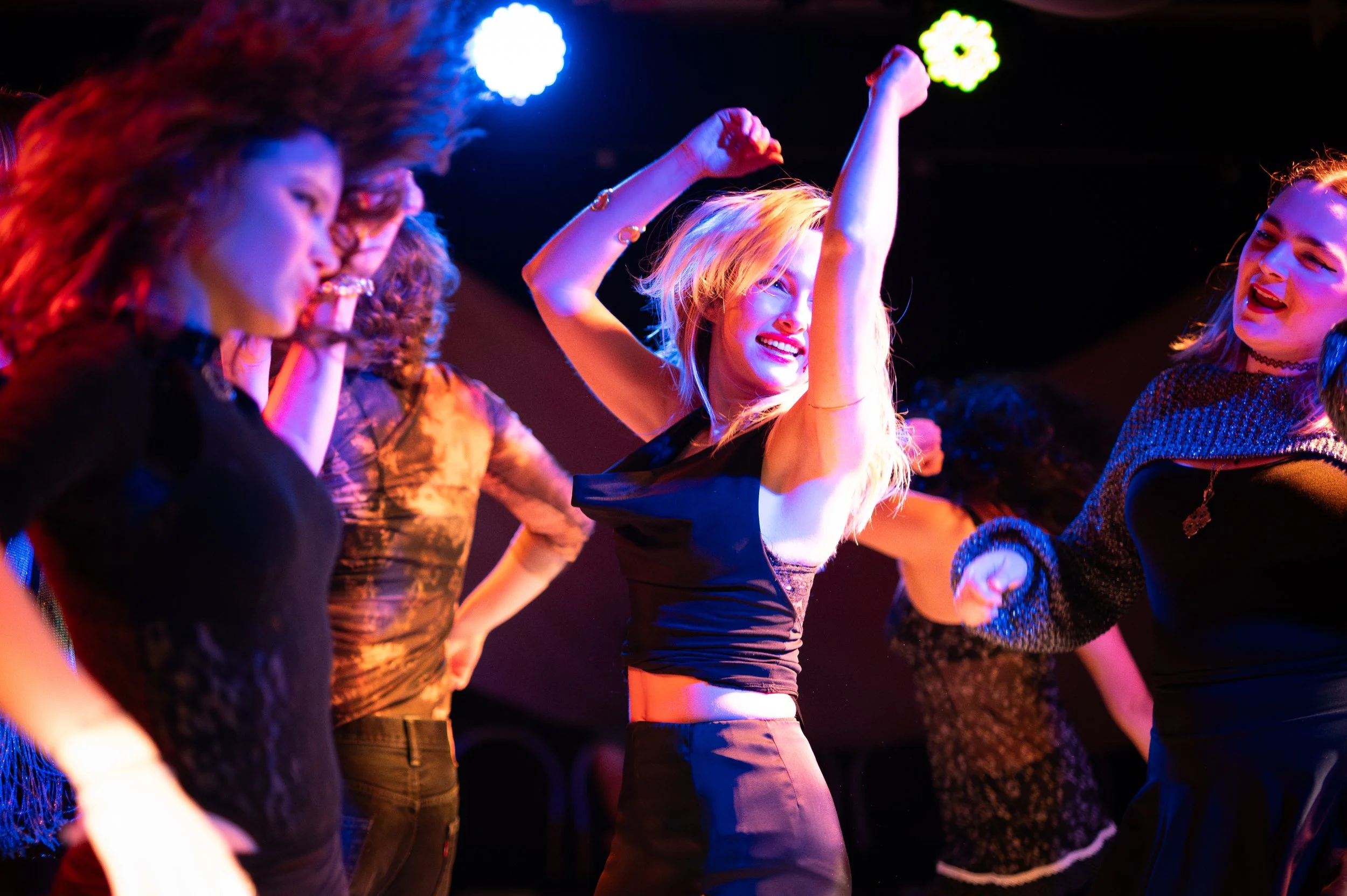 A group of young women dancing together at a party or nightclub, with colorful stage lighting illuminating the scene.