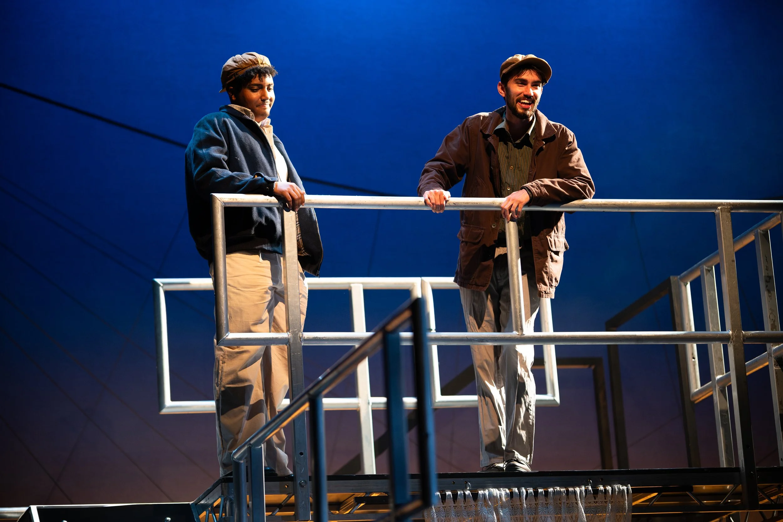 Two men are standing on a metal platform against a dark blue background, engaging in conversation.