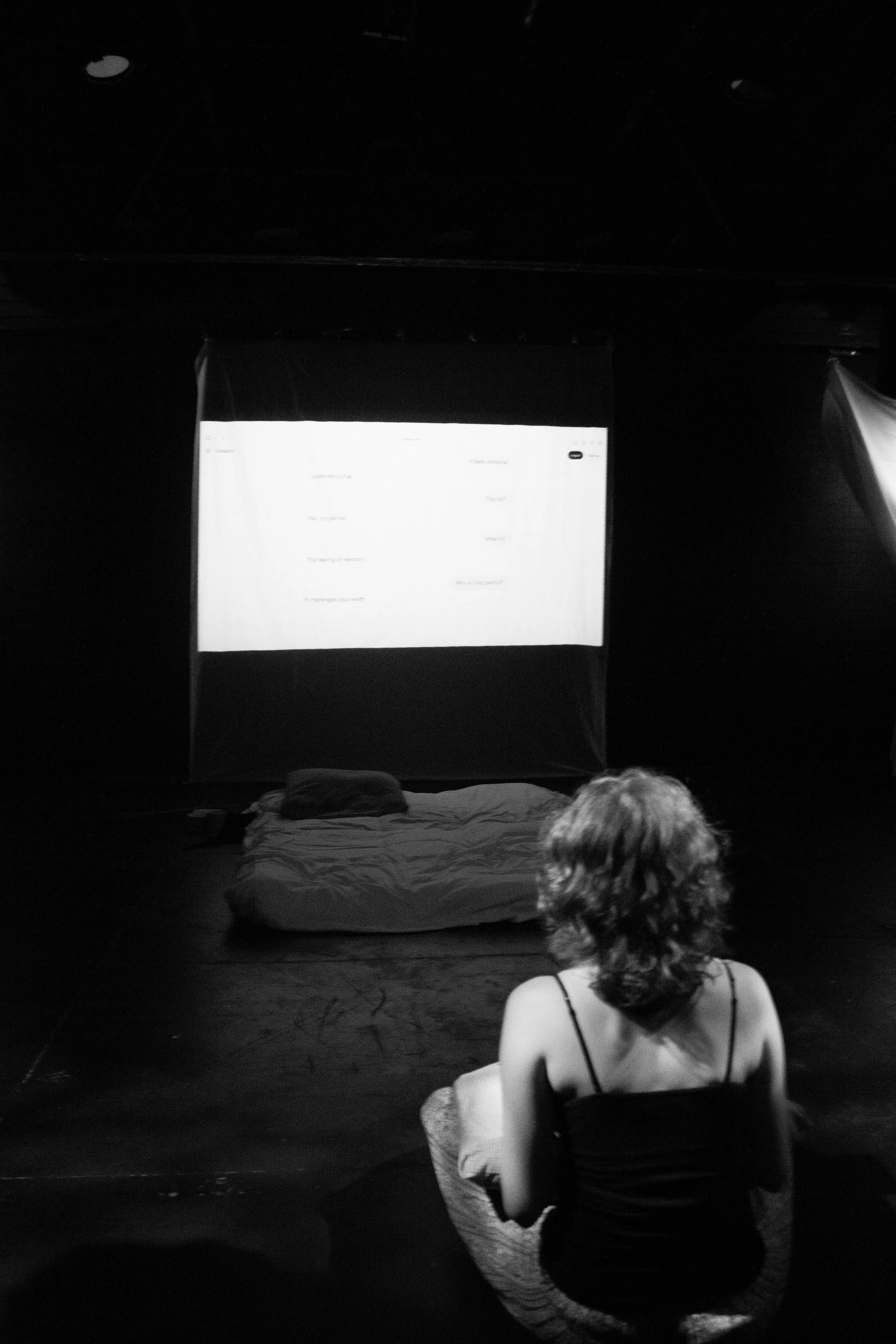 A woman with short curly hair, wearing a black tank top, sitting cross-legged on the floor, facing a large screen with text, in a dark room with a few lights on the ceiling.