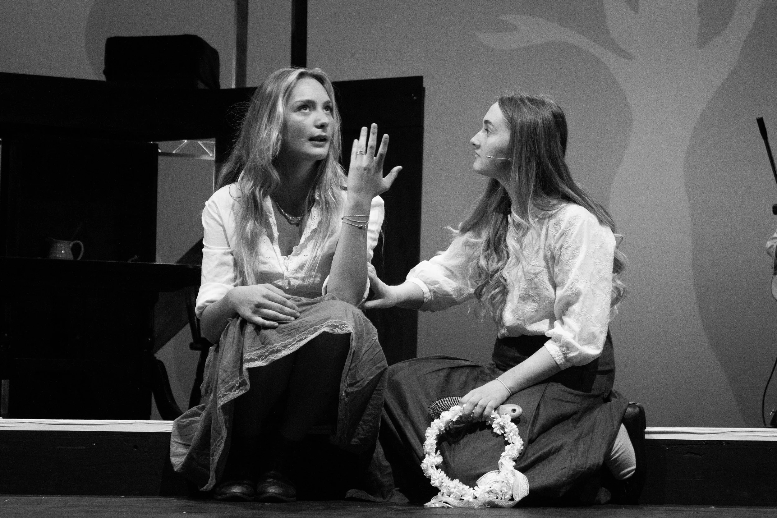 Two women with long hair, one with a microphone headset, sitting close, engaged in a conversation on a stage with a piano and floral decorations in front of them.
