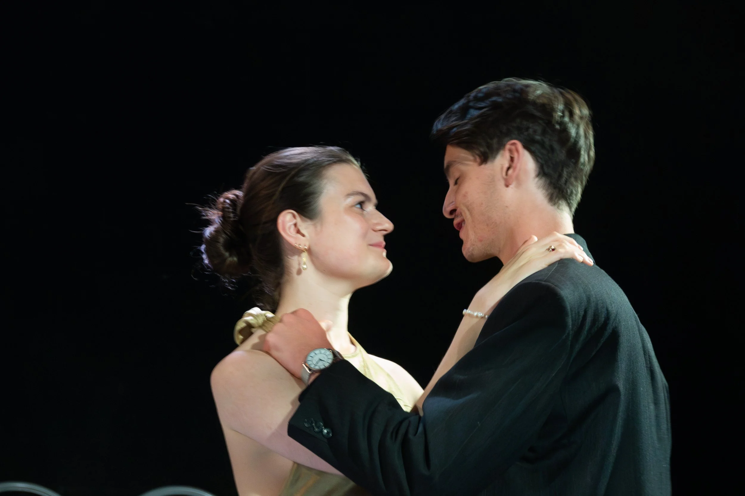 A man and woman dance closely together against a black background, gazing into each other's eyes with smiles.