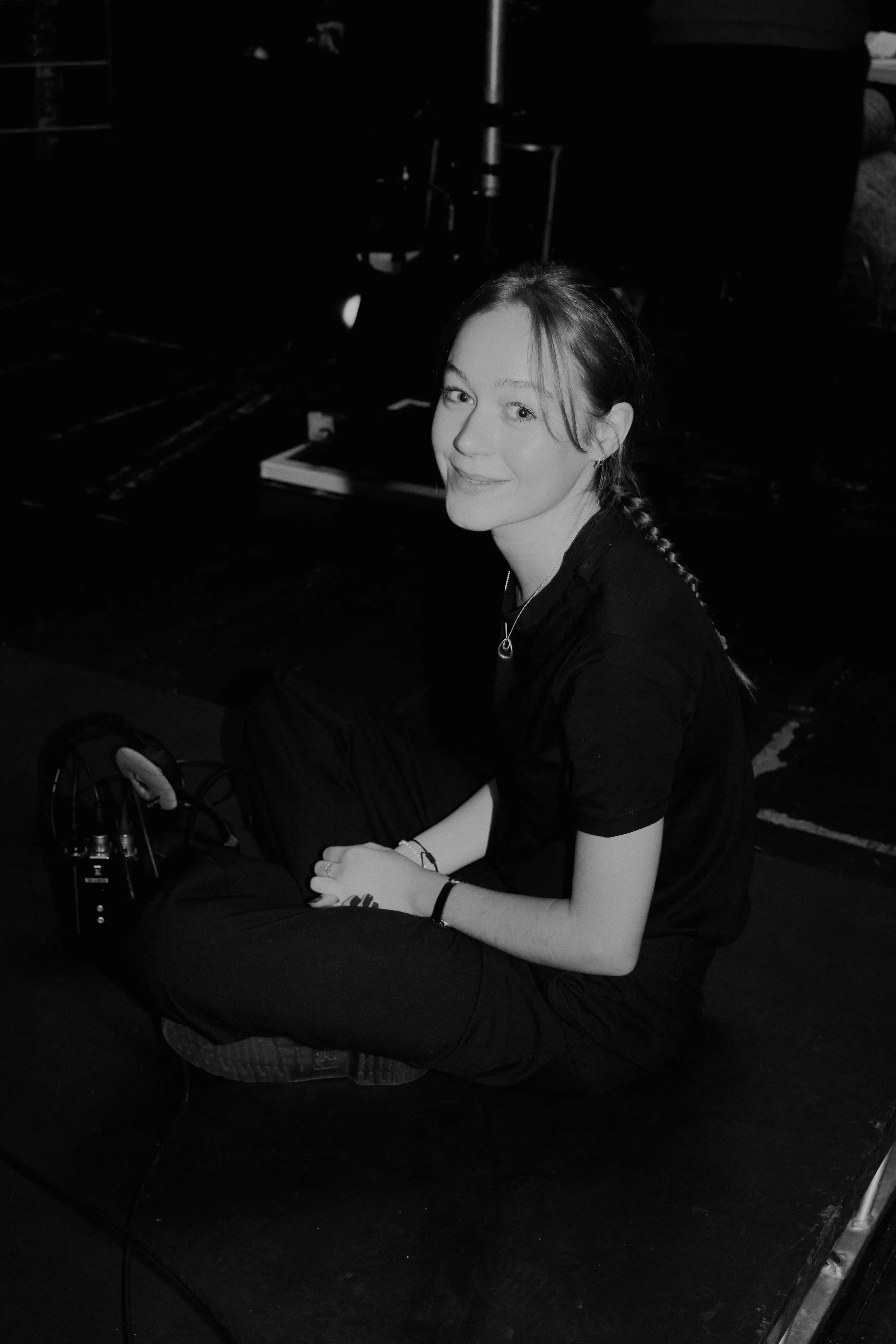 A young woman with long hair in a braid, wearing a black shirt, sitting on the floor, smiling, with some electronic equipment and books around her in a dark room.