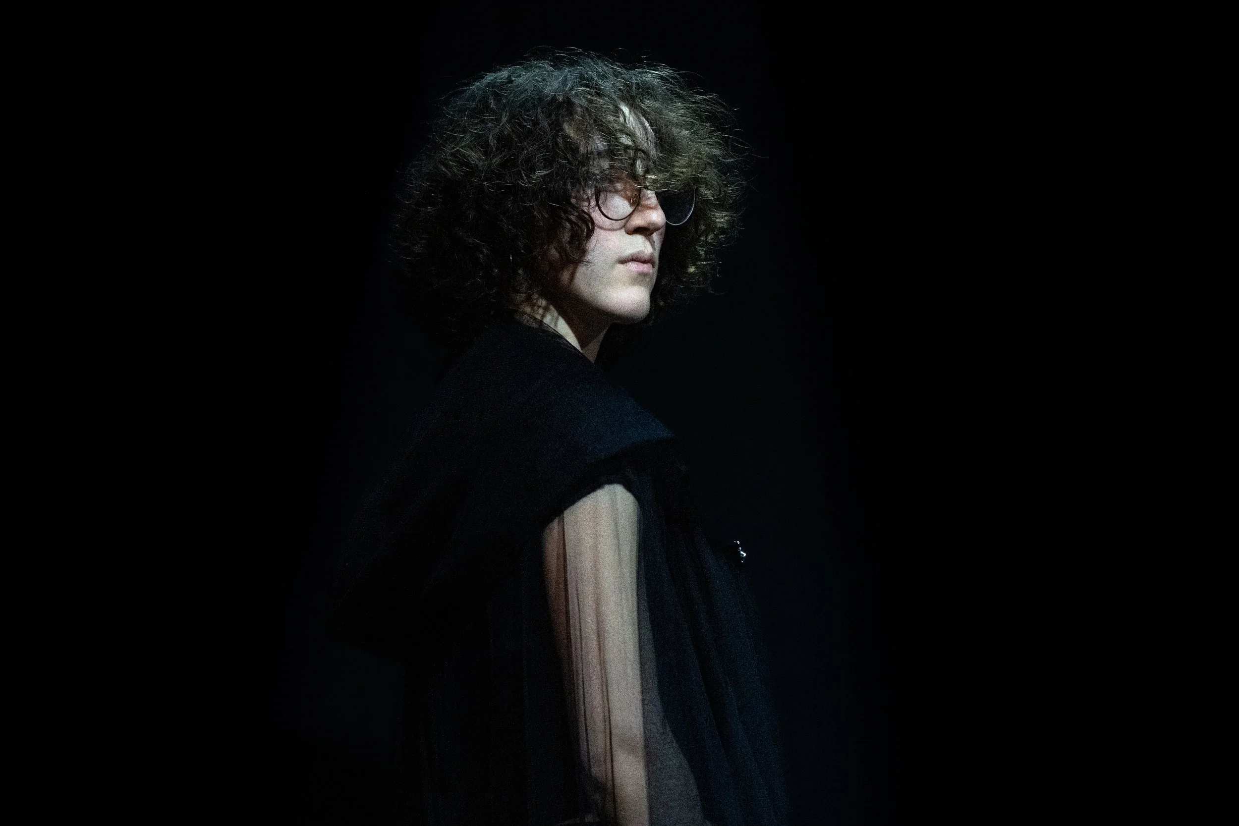 A person with curly hair and glasses, wearing dark clothing, looking to the side against a plain dark background.