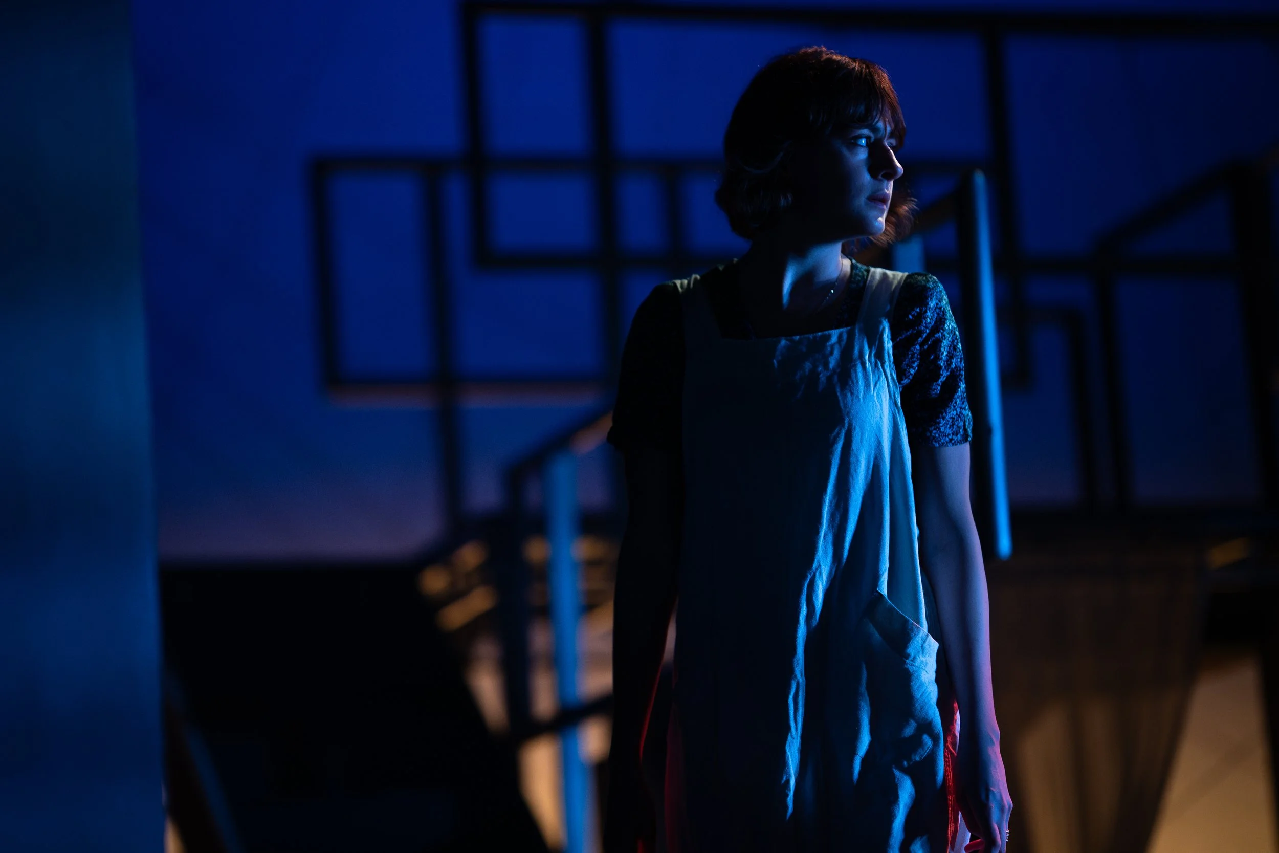 A woman with short brown hair is standing in a dimly lit room with blue and purple lighting, looking to her left. She is wearing a white apron over a dark patterned shirt.