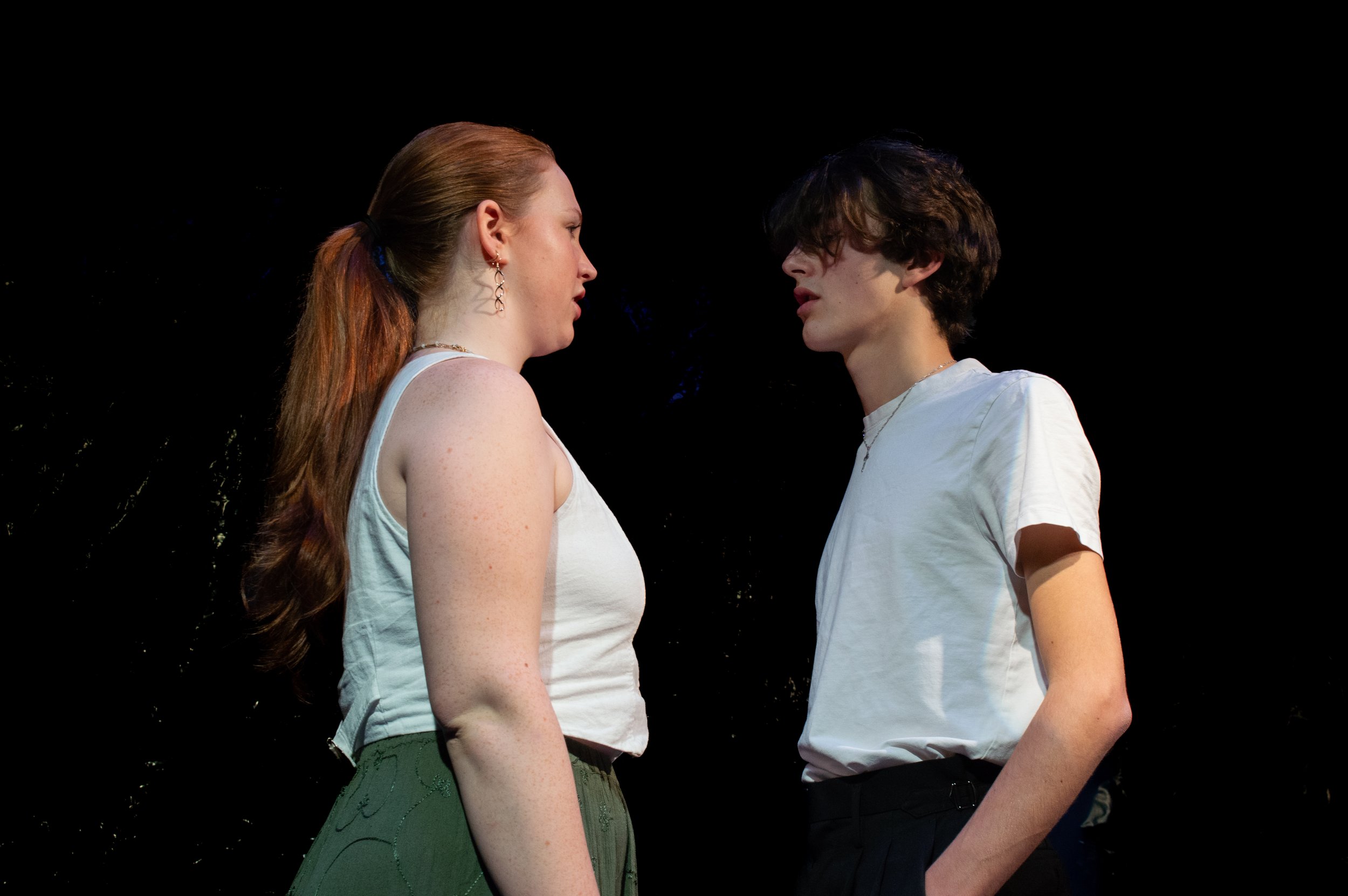 Two young people, a woman with long red hair and a man with curly dark hair, stand close facing each other in a dark background, seemingly engaged in an intense moment.