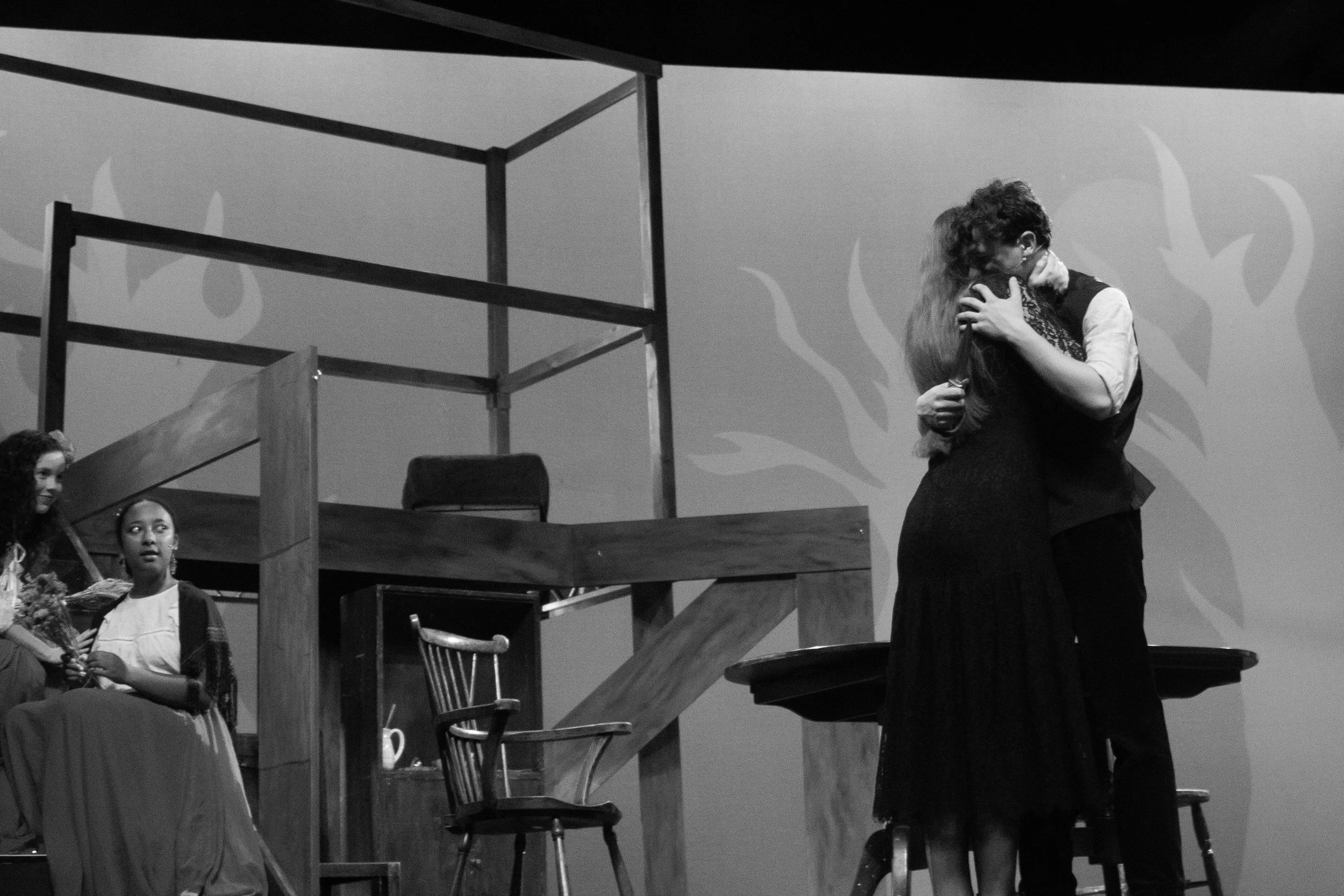Two women hugging on stage during a performance or event, with a background wall and stage furniture including chairs and a wooden structure.