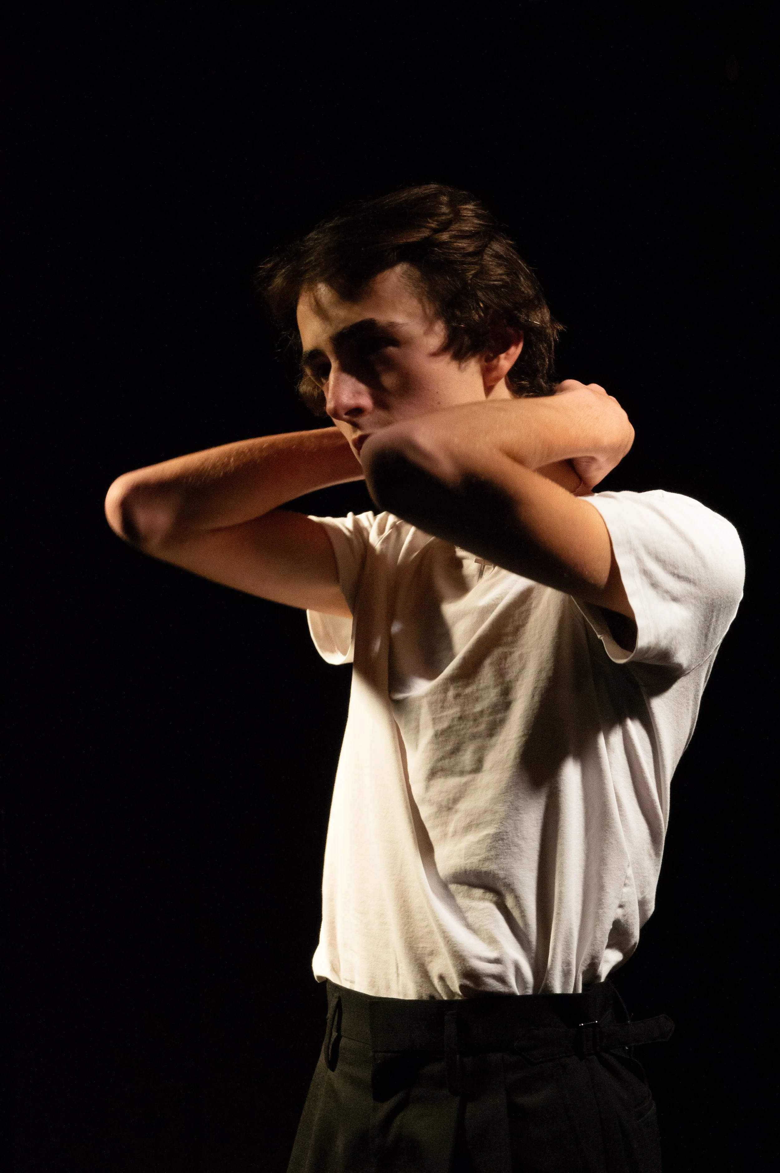 A young man with medium-length dark hair wearing a white t-shirt and black pants stands in a dark environment, with his arms crossed behind his head.