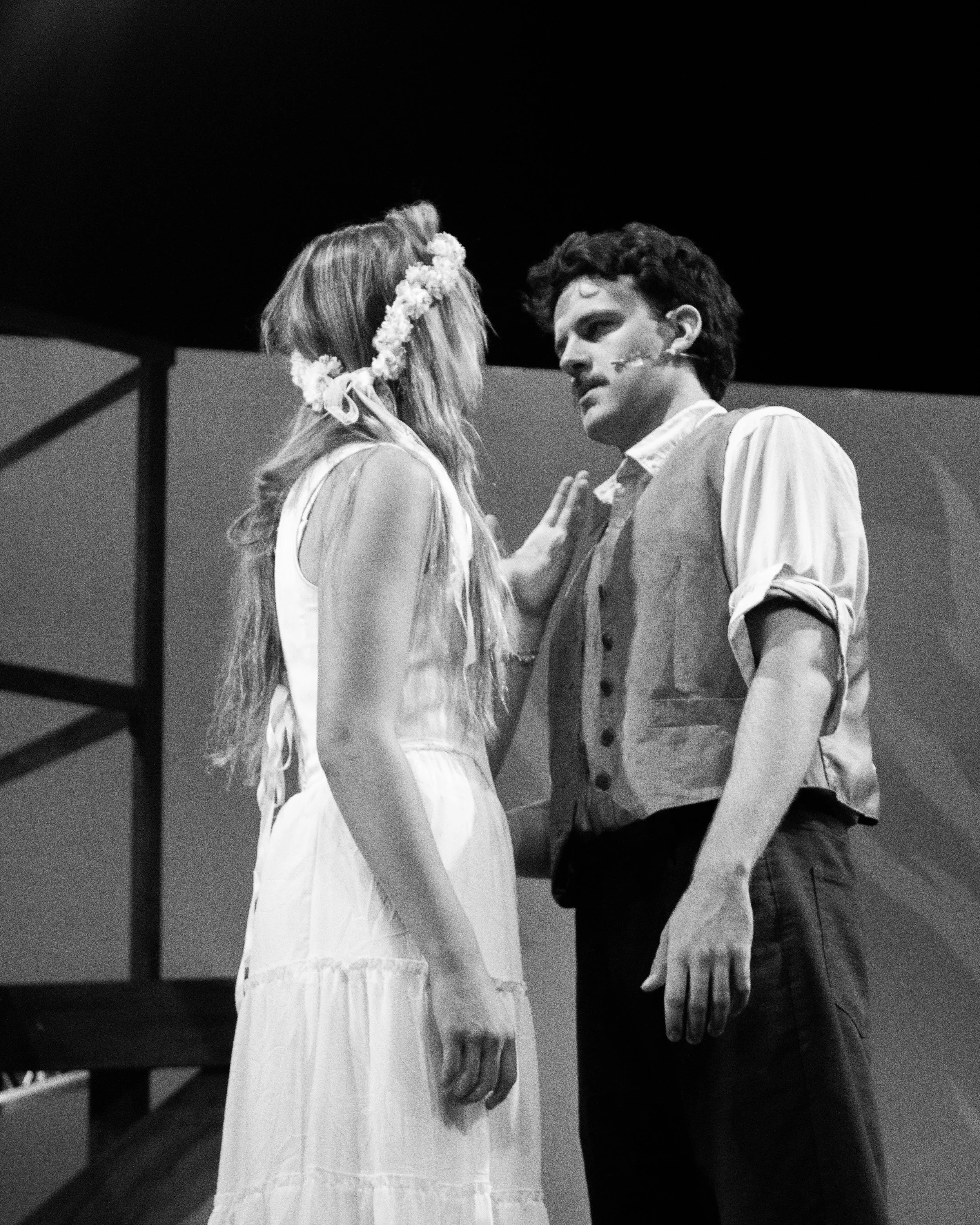 A black and white photo of a man and woman facing each other closely, holding hands. The woman wears a flower crown and a light dress, while the man wears a shirt with rolled-up sleeves and a vest. They appear to be on stage, possibly in a theatrical performance.
