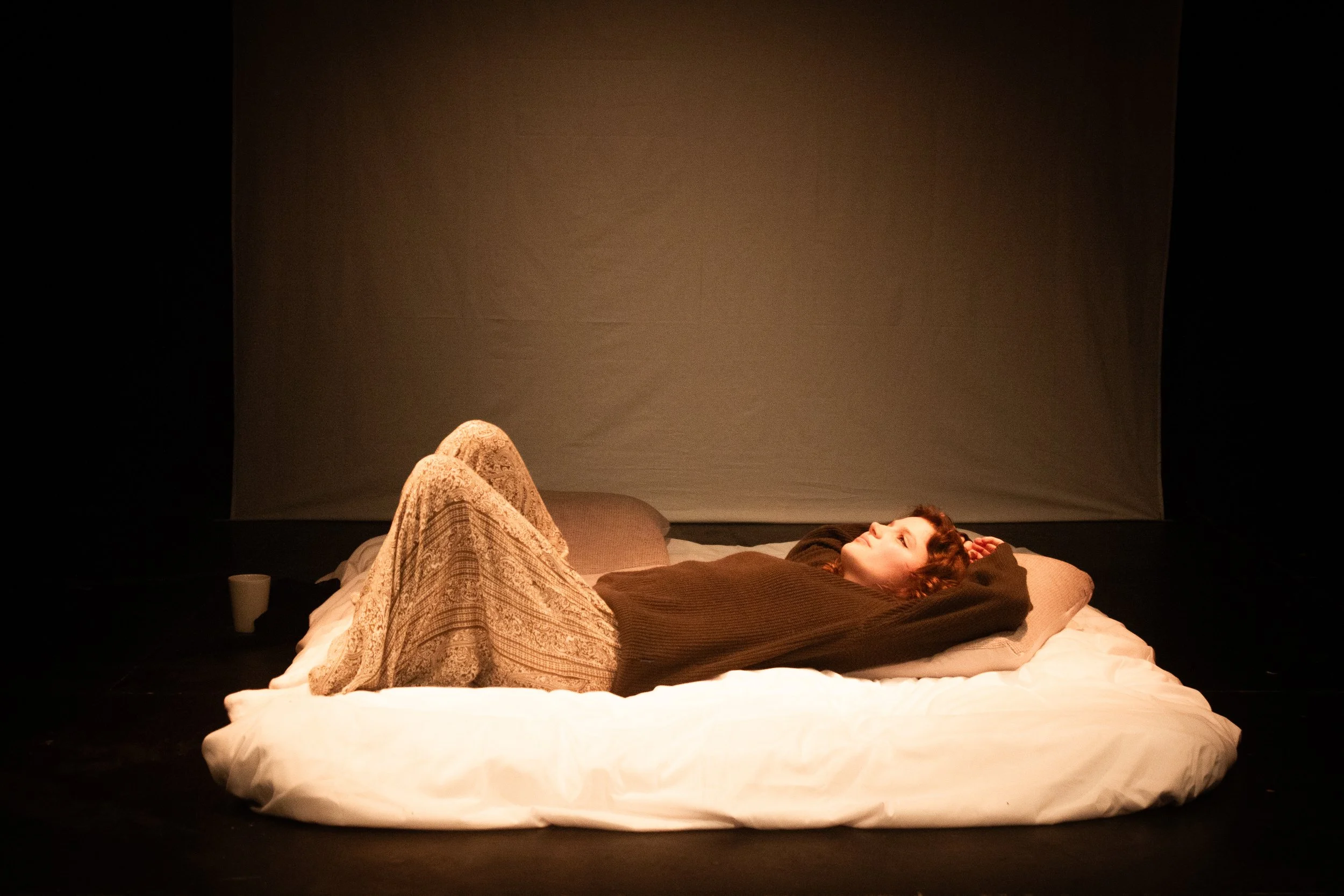 A person with curly hair lying on a bed, resting their head on a pillow, with their eyes closed, set against a plain, dark background.
