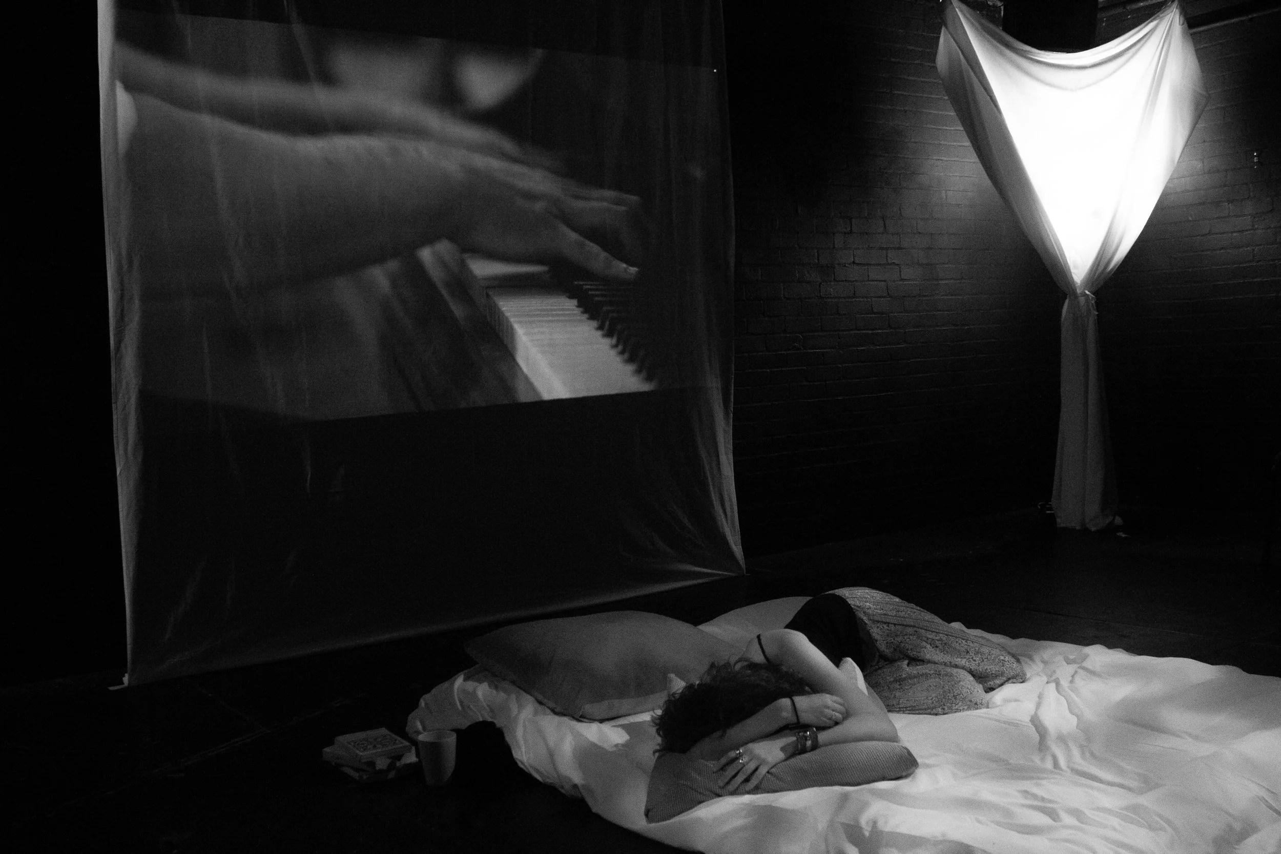 A person lying on a bed in a dimly lit room, with a projection of a hand playing piano on the wall and a fabric draped in a curtain-like manner.
