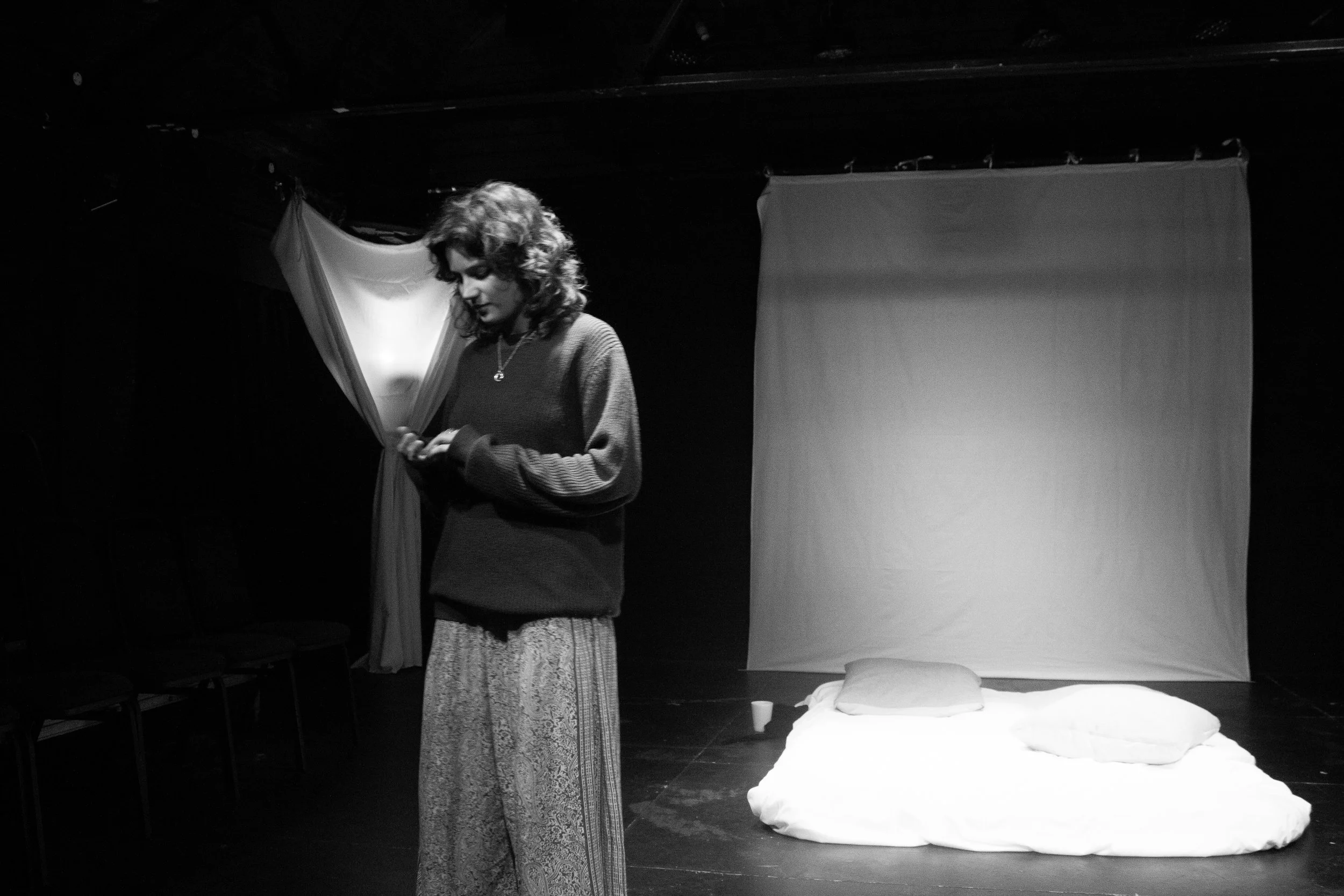 A woman with curly hair standing in a dark room, looking down at her phone. Behind her are white curtains and a bed with pillows and white bedding. The setting appears to be a performance or rehearsing space.
