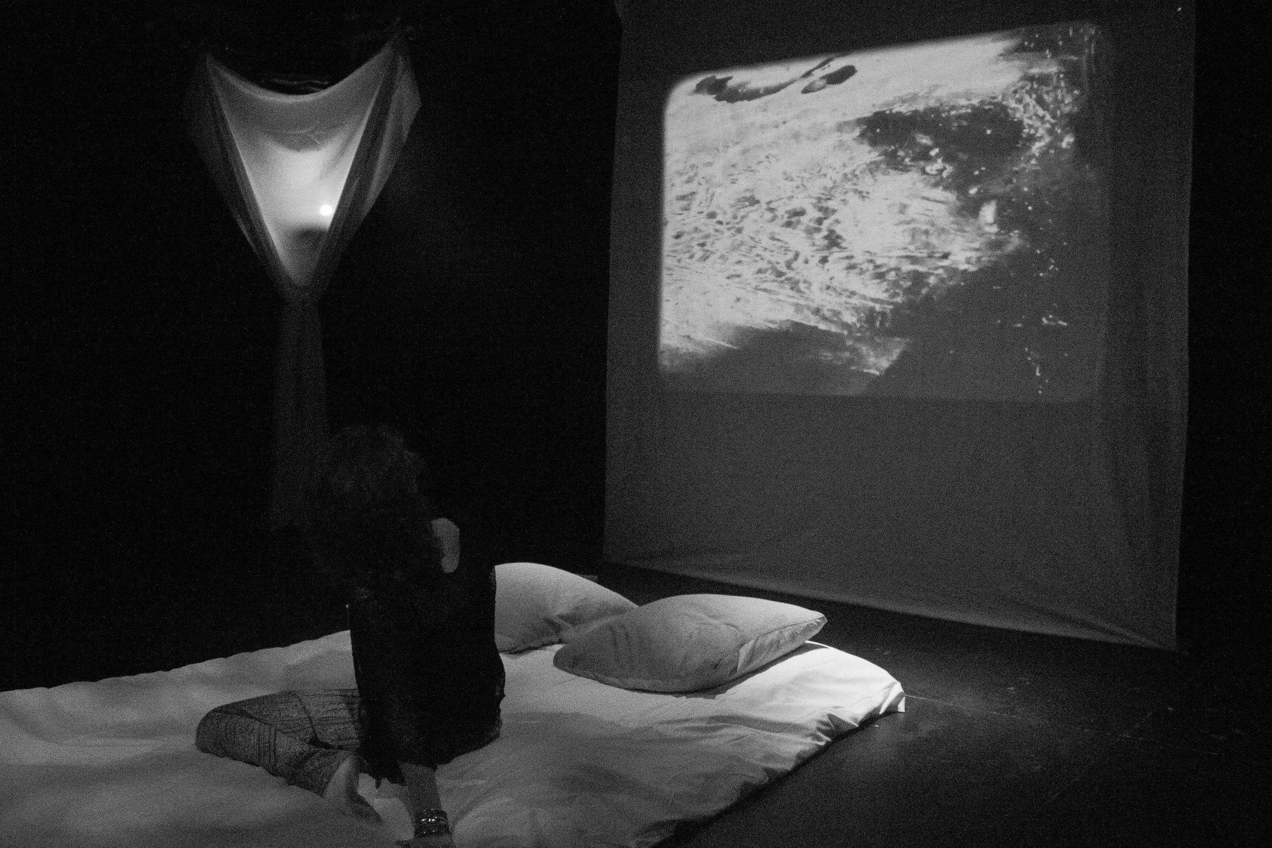 Black and white photo of a person wearing a dark jacket, striped pants, and boots, lying on a bed with pillow and blanket, facing towards a window showing a night sky with stars.
