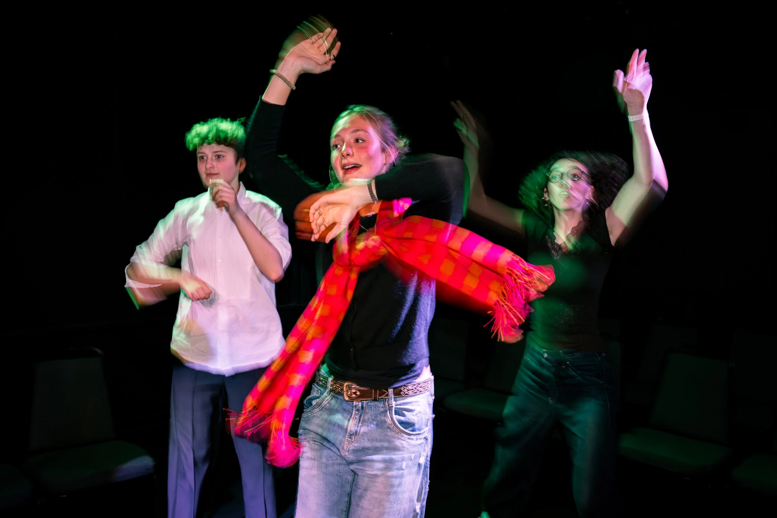 Three young women dancing under colorful stage lights against a dark background, with their hands raised and expressive faces.