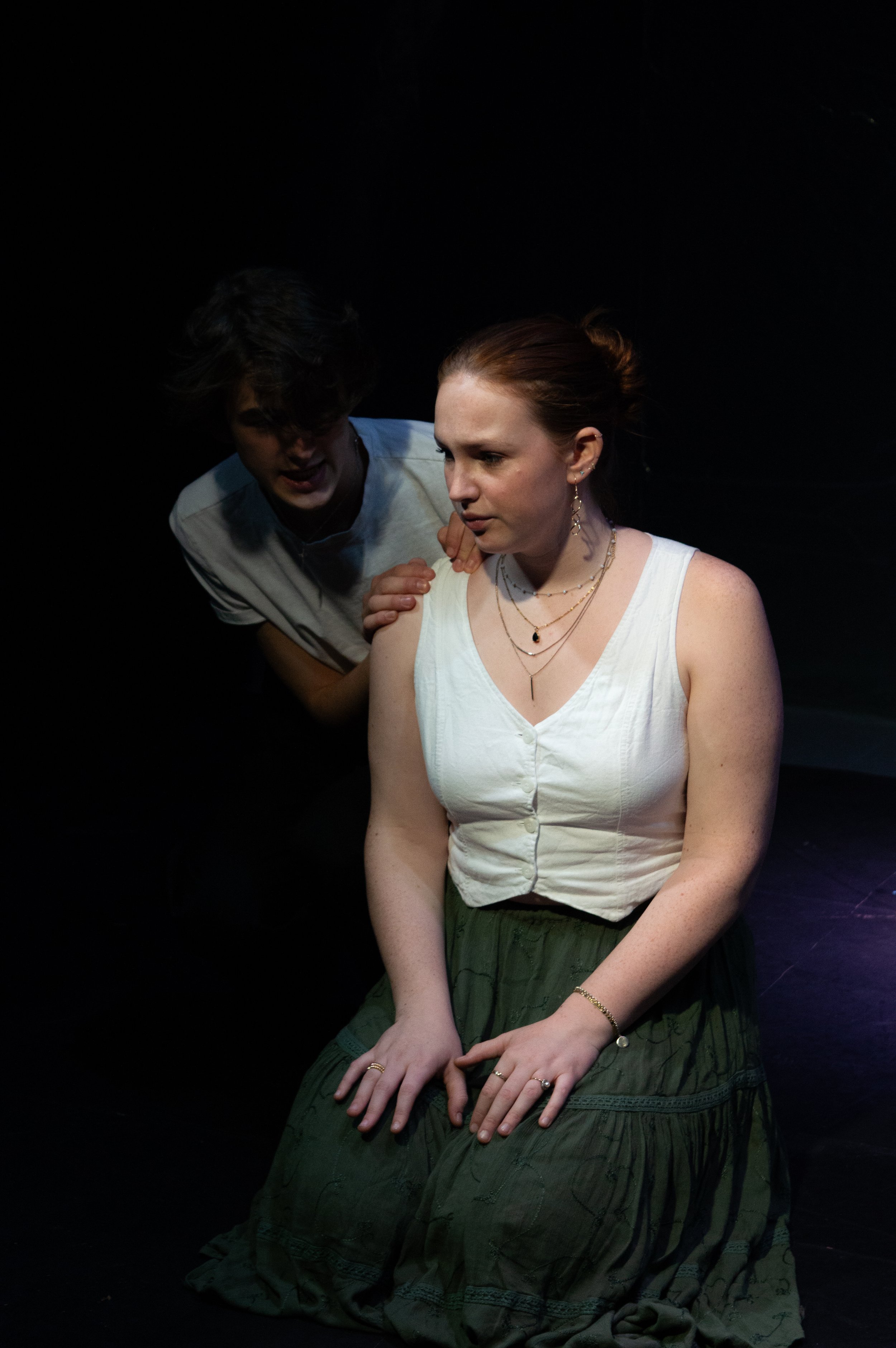 A woman with red hair sitting on a dark stage with her hands resting on her lap, wearing layered jewelry, a white sleeveless top, and a green skirt, while a man with dark hair, wearing a white shirt, leans in close behind her, appearing to speak to h