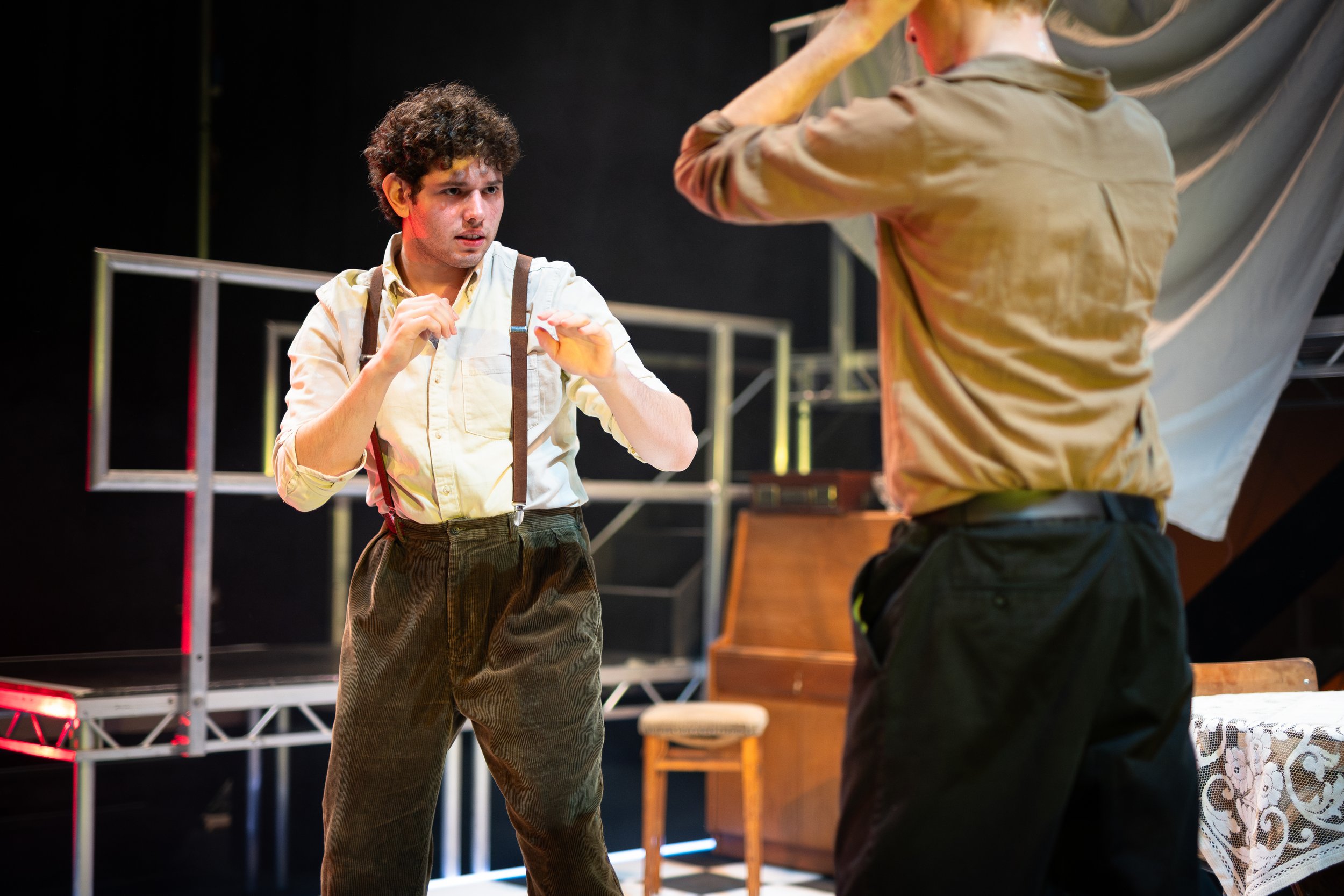 Two actors on a stage engaged in a theatrical scene, with one person in a white shirt and suspenders looking at another person in a tan shirt. The background includes stage equipment and a draped fabric.