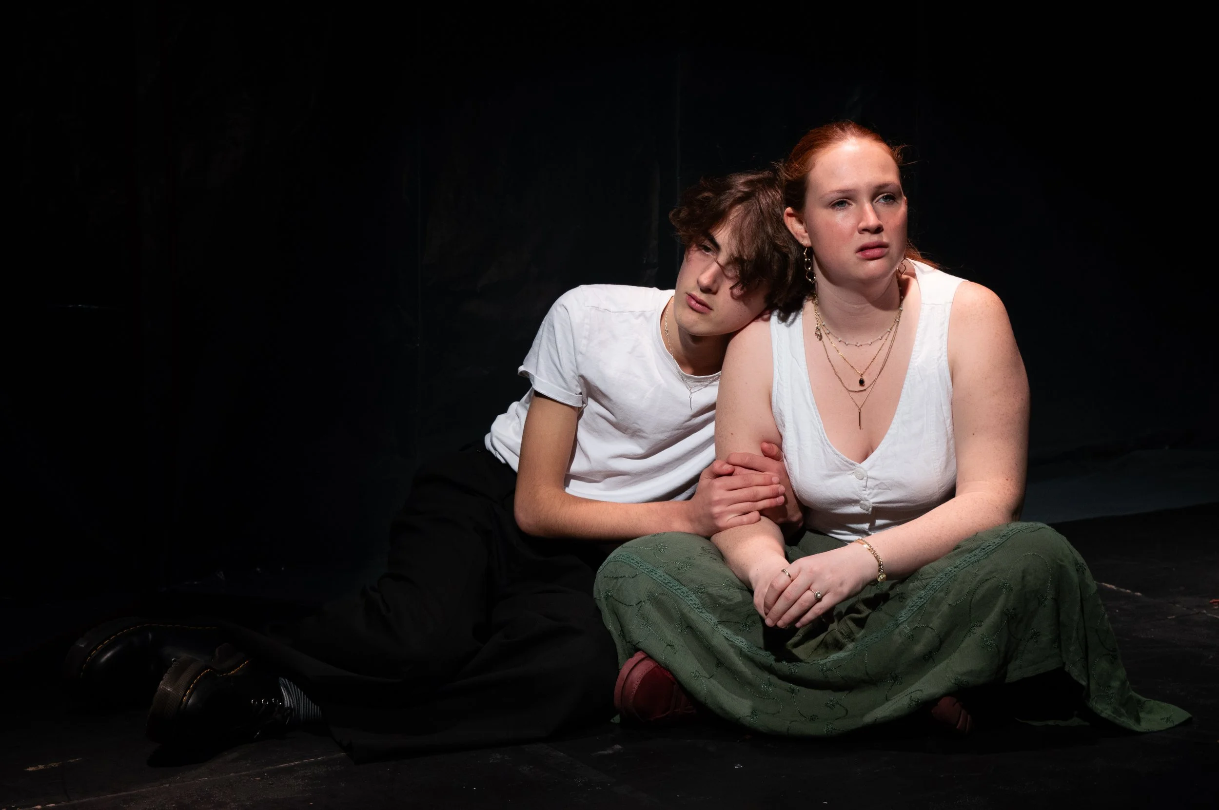 Two young women sitting together on a dark stage; one with red hair and a white sleeveless top, the other with short curly hair and a white t-shirt, showing an emotional moment.