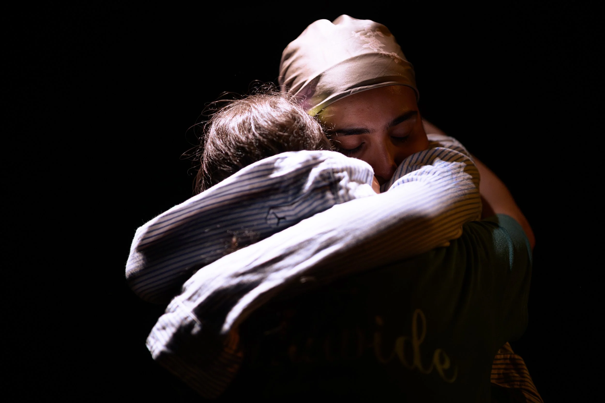 A person wearing a headscarf is hugging a child with curly hair in a close embrace, with both individuals having their eyes closed, against a dark background.