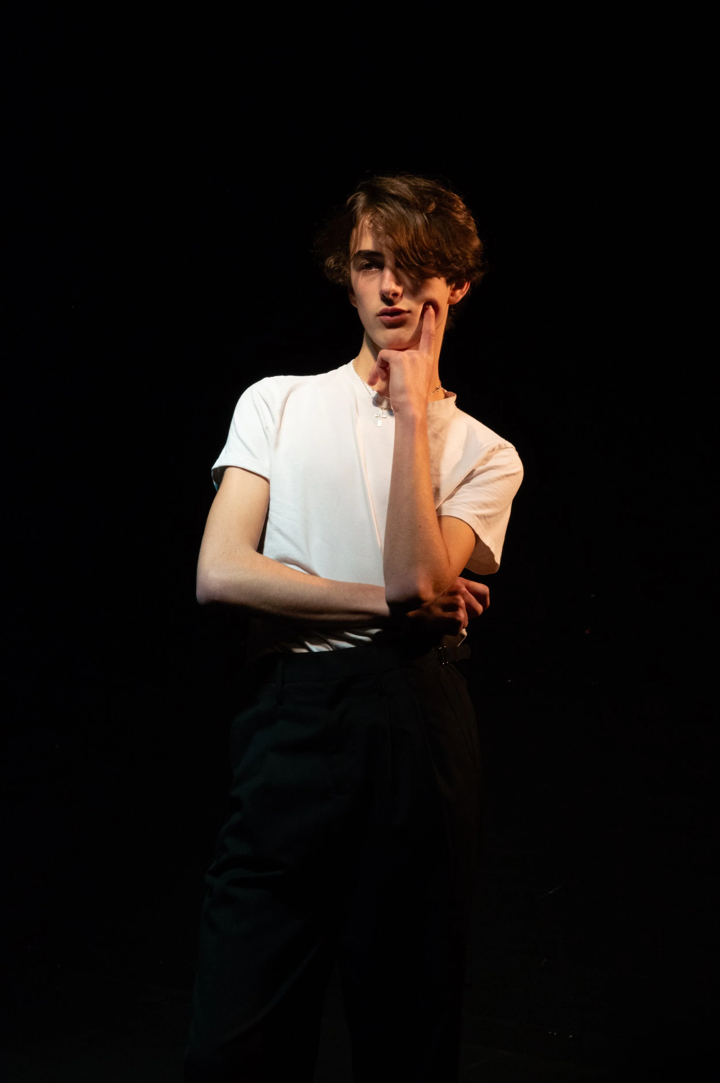 A young woman with short wavy brown hair and light skin posing against a black background, wearing a white t-shirt and black pants, with one hand resting on her chin and the other crossed over her stomach.