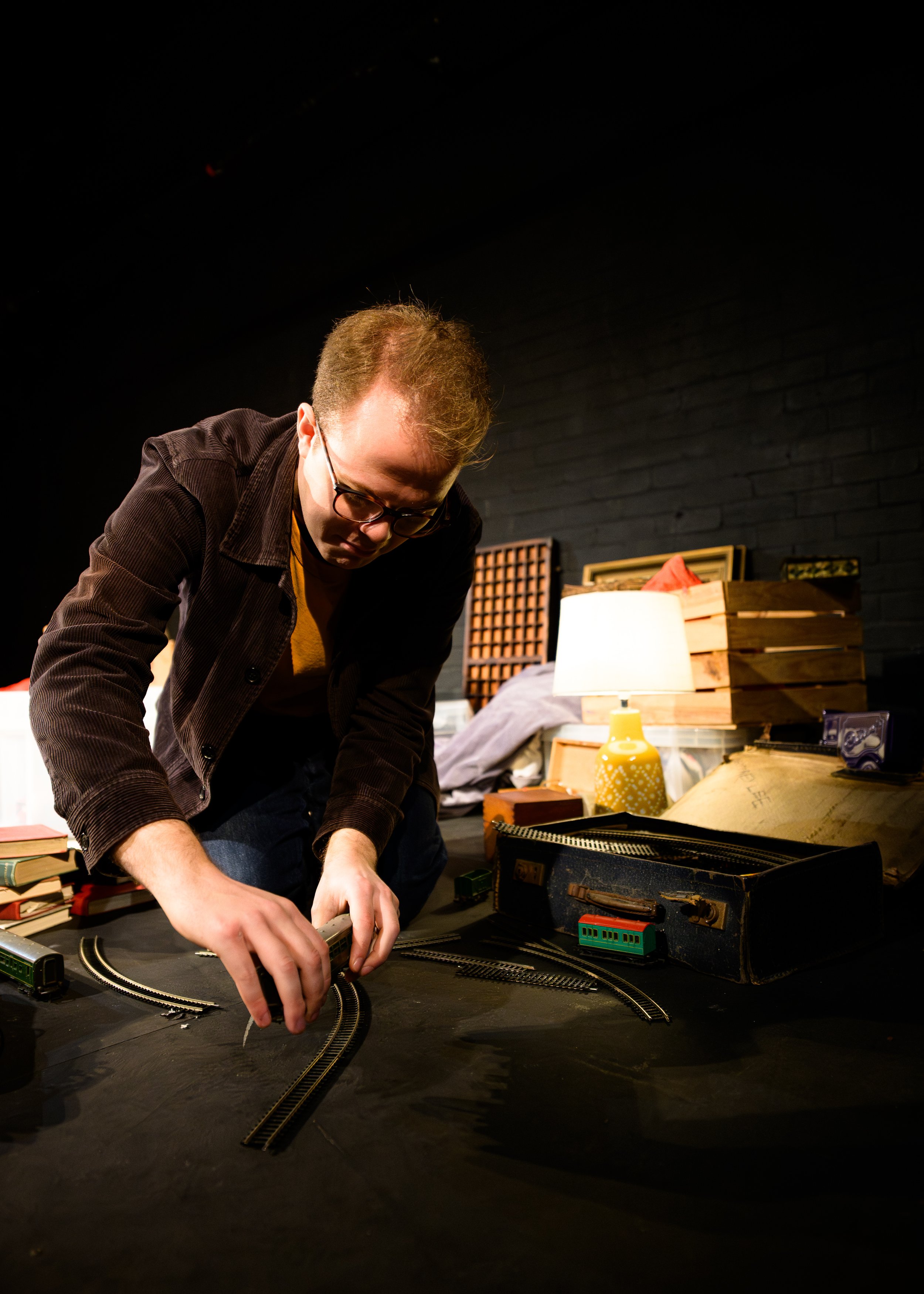 A man with glasses arranges model train tracks on a table, with various boxes and items around him, in a dimly lit room with a black brick wall.