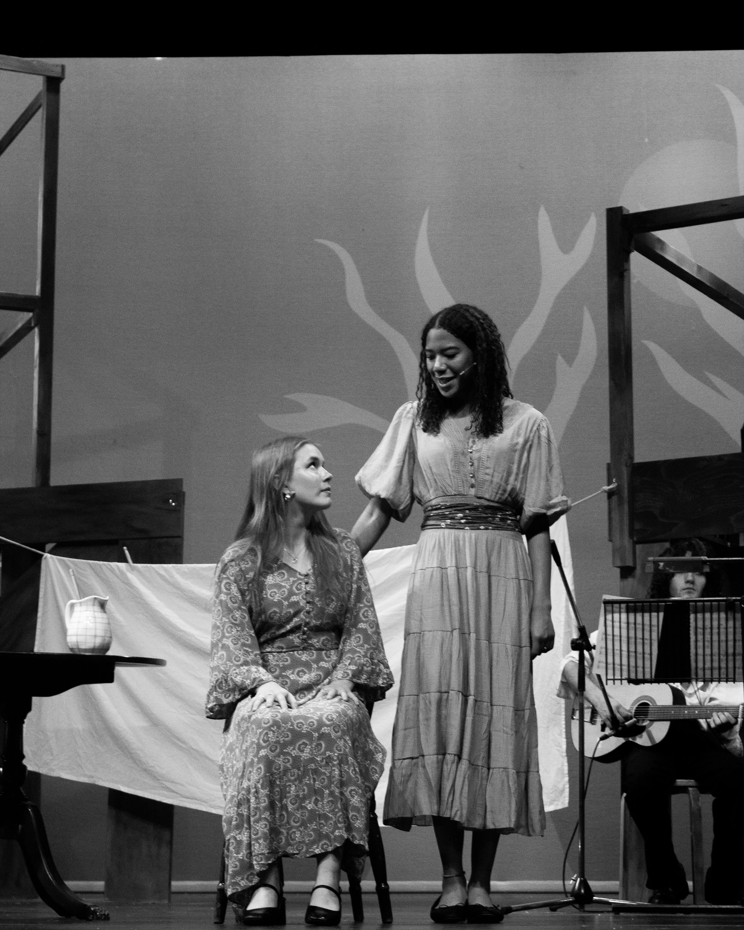 Two women in vintage clothing on a stage, one sitting and the other standing, with musical instruments and stage props around them.