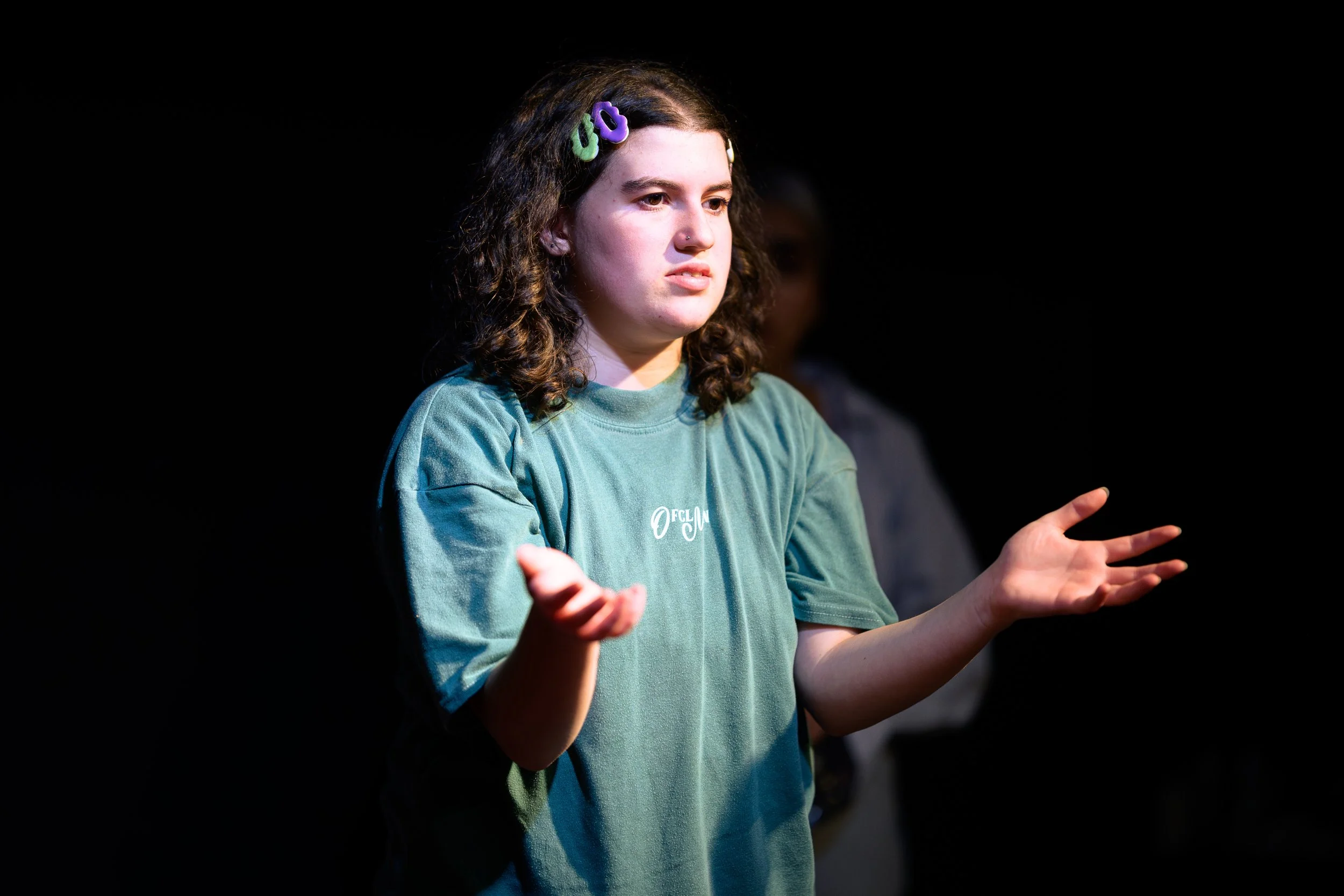 A young woman with curly brown hair wearing a green t-shirt with a logo, speaking on stage with a black background.
