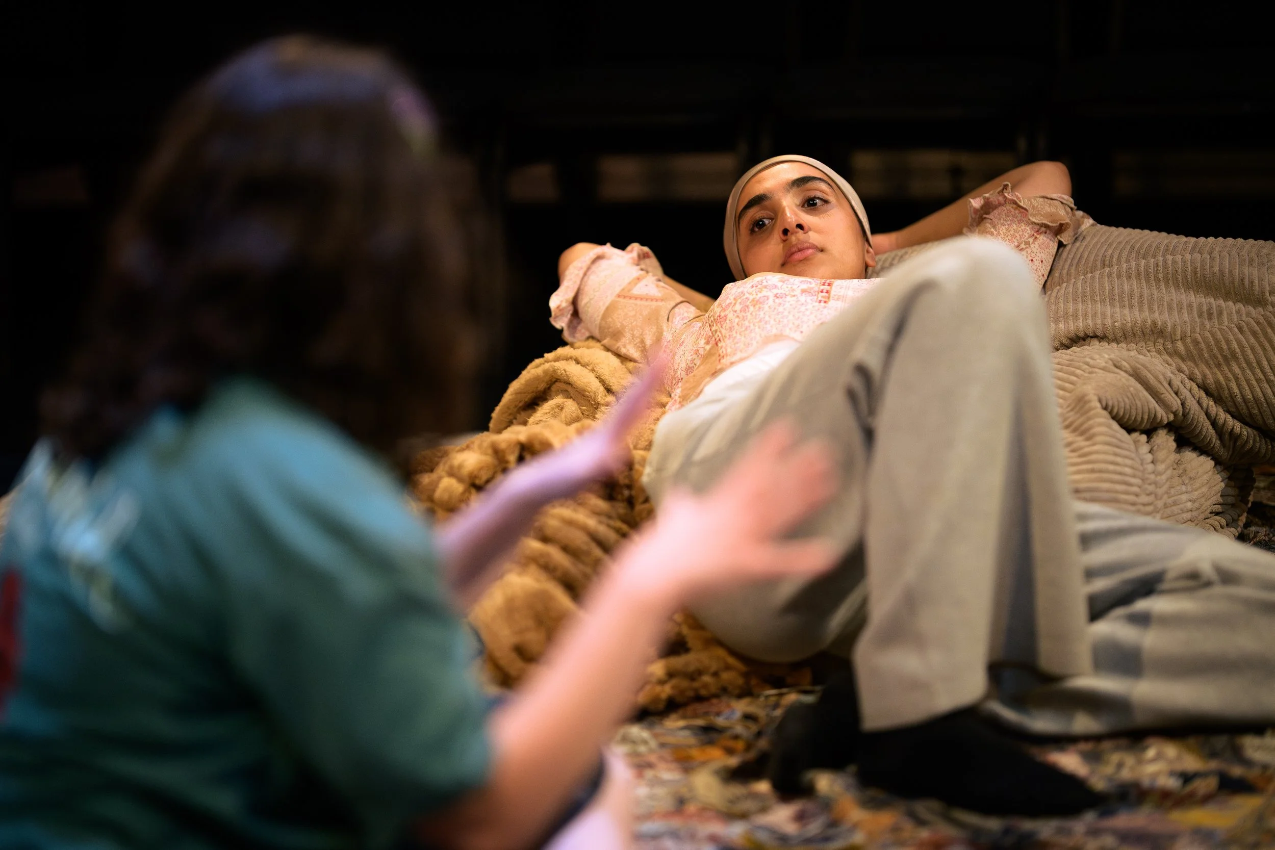 A woman lying on her back on a bed, looking at a person in the foreground who is holding a phone. The woman is wearing a headband and a floral top, with blankets and pillows around her.