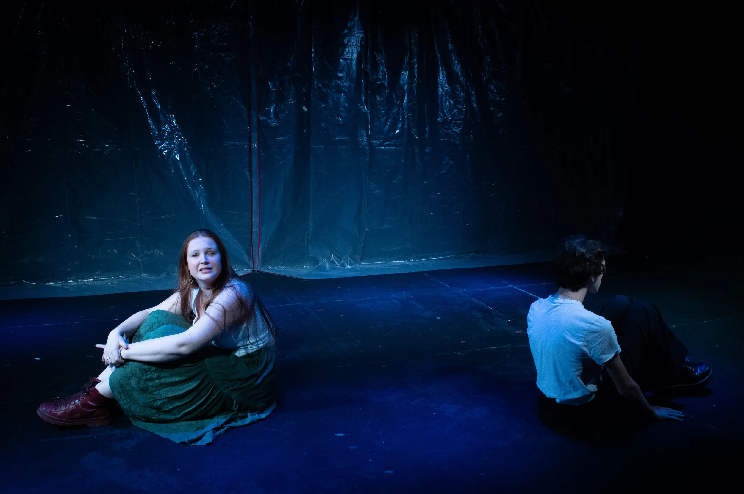 Young woman with red hair sitting on stage, wearing a green dress and red boots, with two other performers sitting nearby, against a dark, textured background.