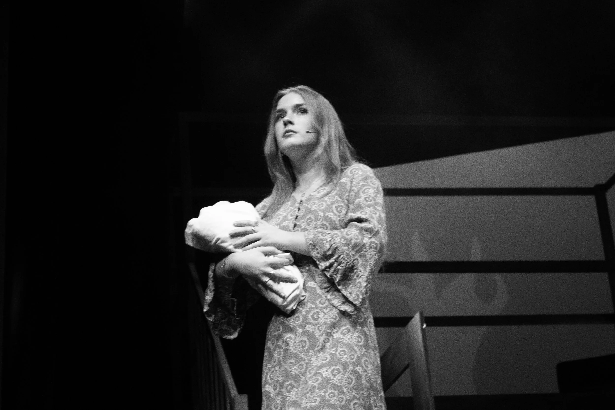 A woman with long hair, wearing a patterned dress, holding a small animal, possibly a puppy, on stage during a presentation or performance.