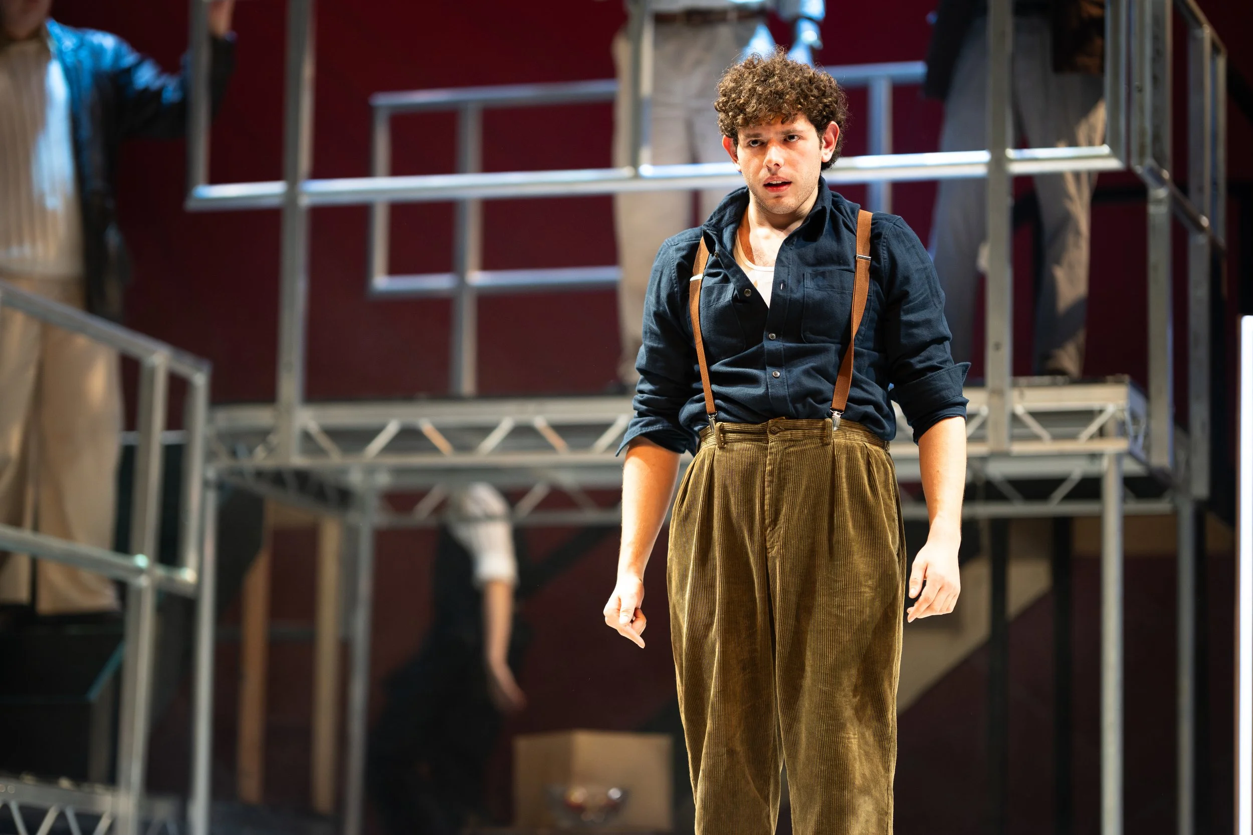 A young man with curly hair, dressed in a dark shirt and brown suspenders, standing in a theater or stage setting, looking confused or concerned.