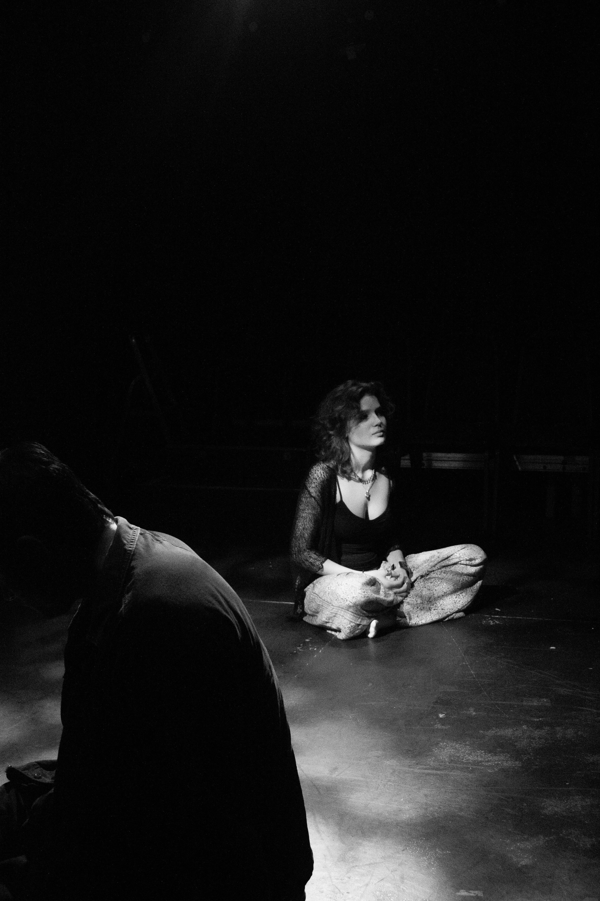 A woman sitting cross-legged on the floor in a dark space, wearing a black top and light-colored patterned pants, with an intense expression. Partially visible is another person in the foreground, facing away.
