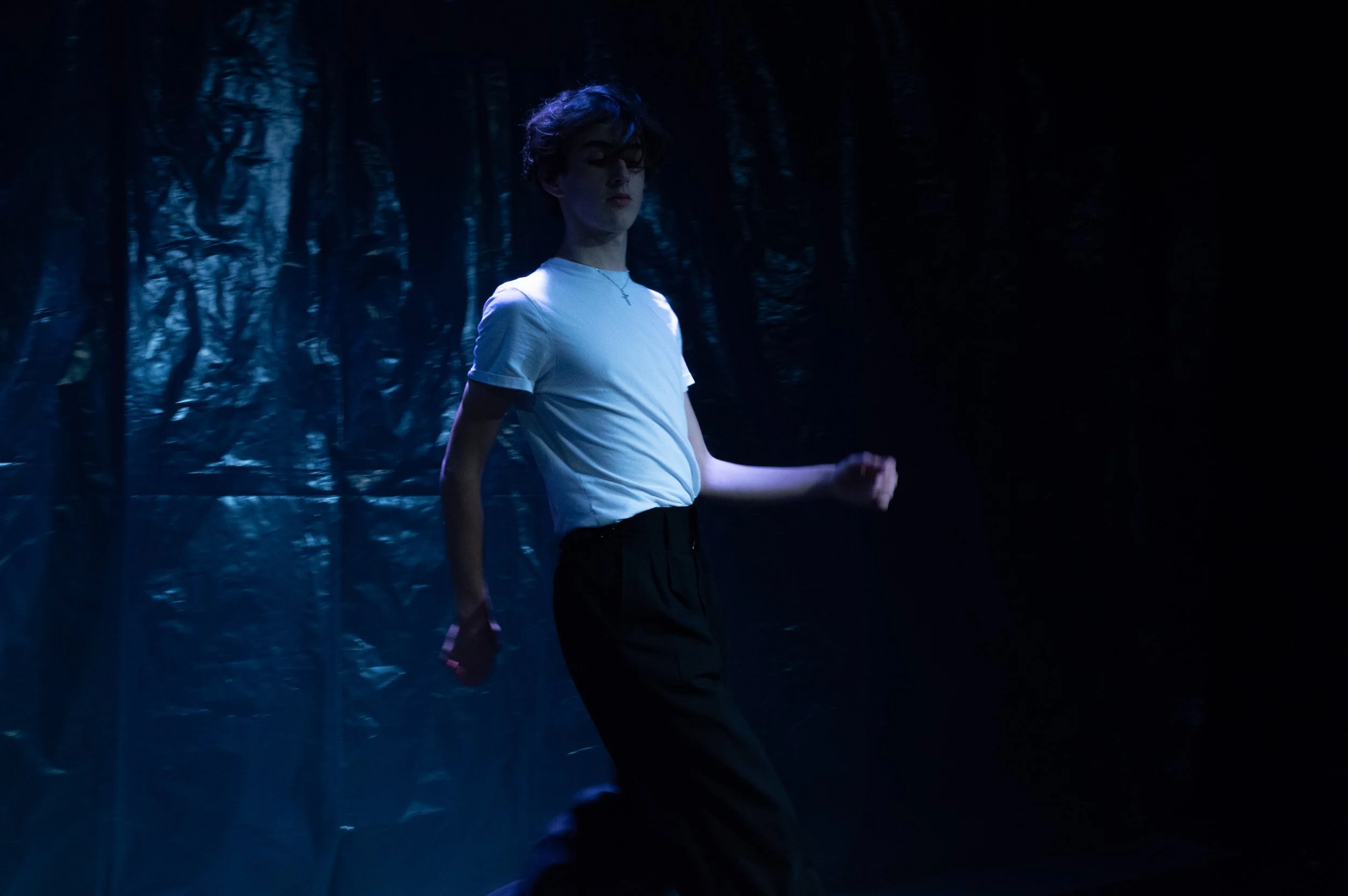 A young person in a white shirt and black pants standing in a dark environment with faint blue lighting, surrounded by dark, textured surfaces.