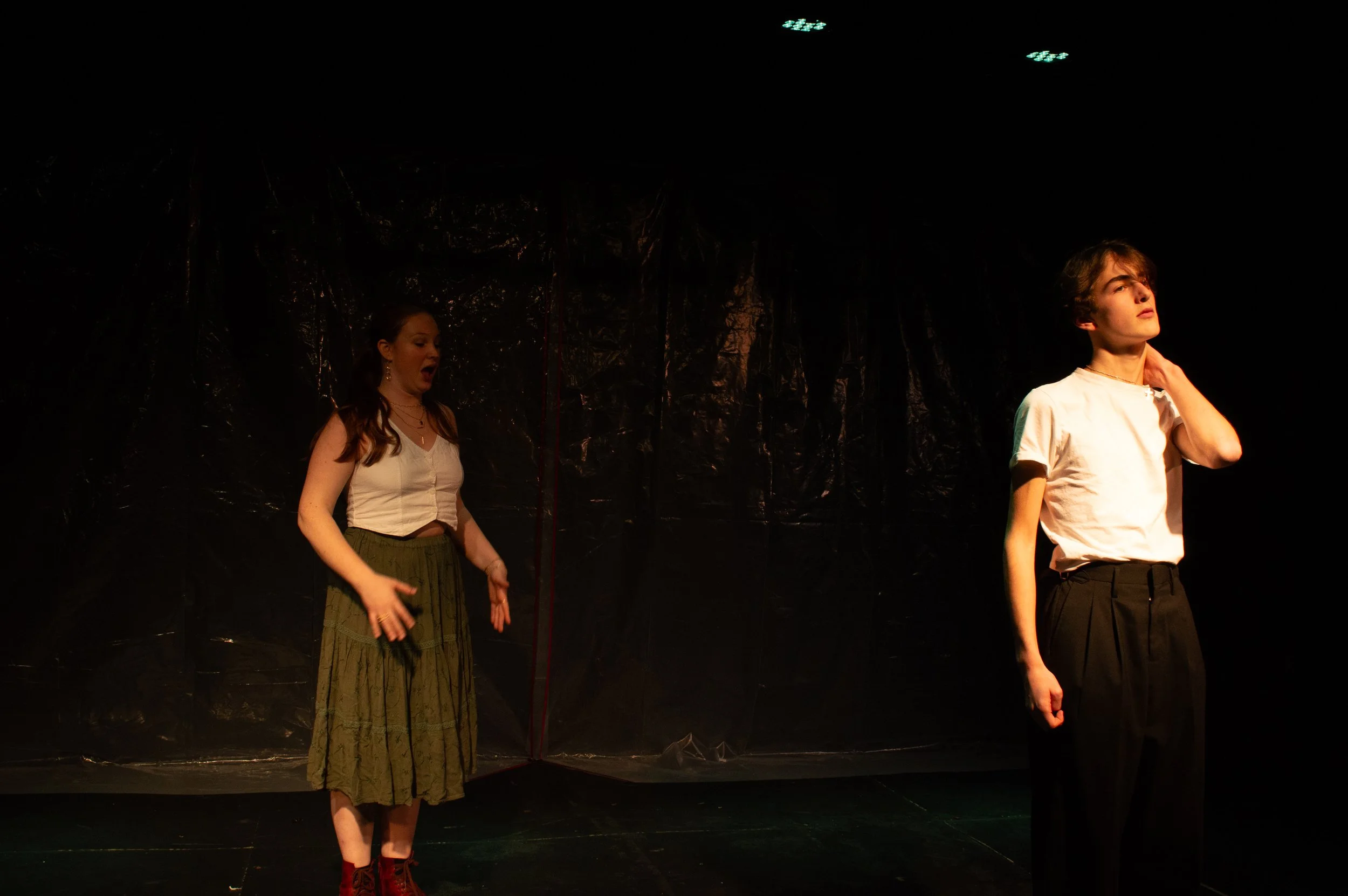 Two young performers on stage during a theatrical performance, with a dark backdrop and overhead stage lights. The girl on the left is wearing a white shirt and a long skirt, and the boy on the right is wearing a white T-shirt and dark pants.