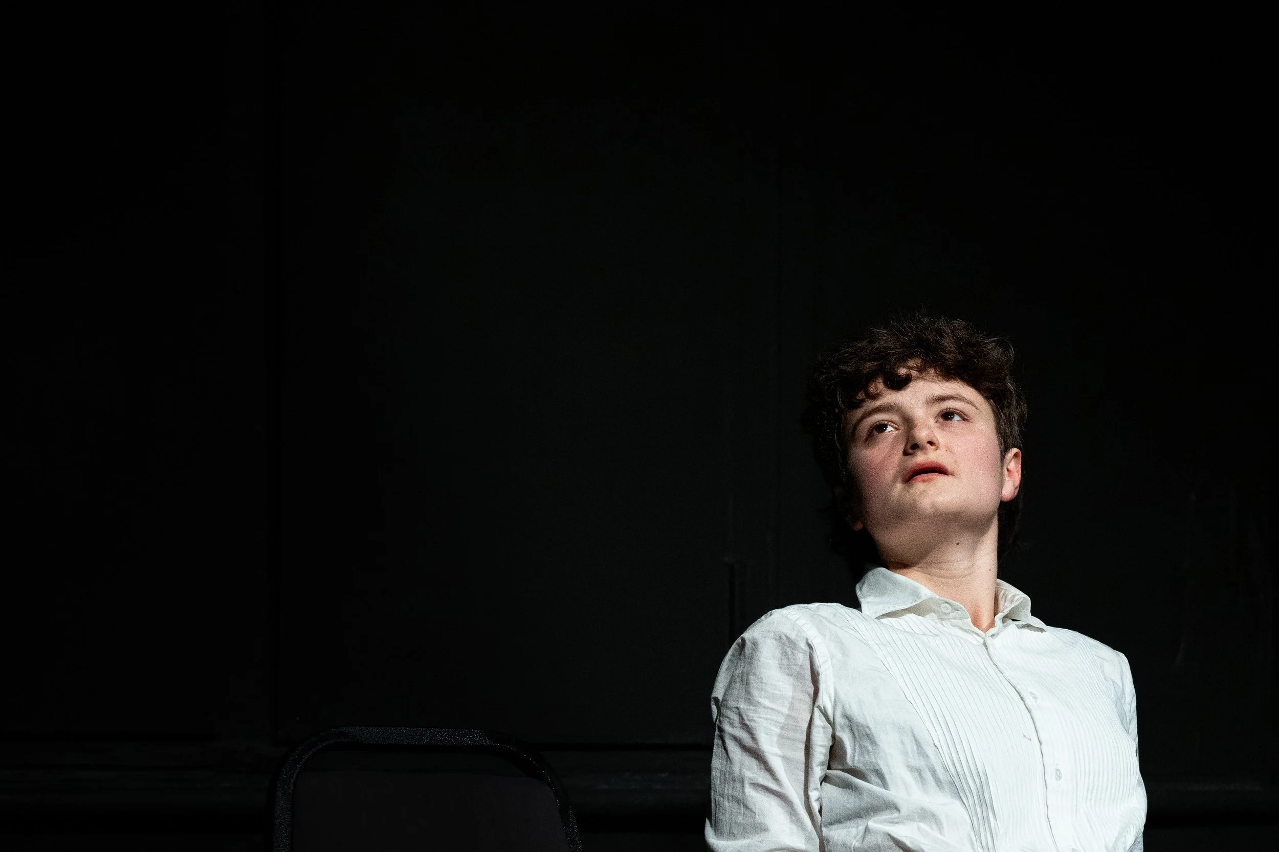 Young boy with curly hair wearing a white button-up shirt, looking upward, against a black background.