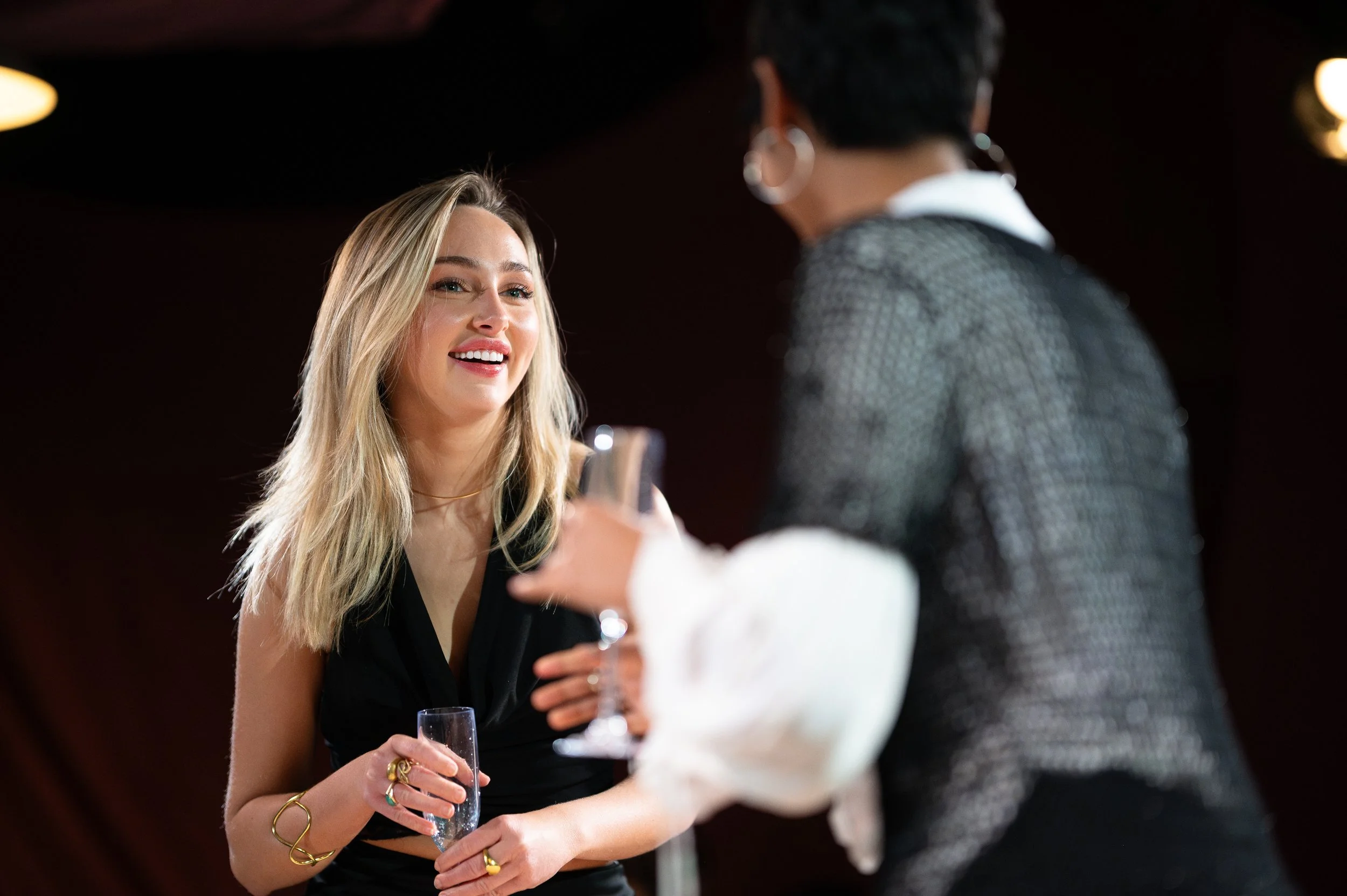 A young woman with long blonde hair and a black dress smiling while holding a champagne glass, engaging in conversation with another person at a social event.