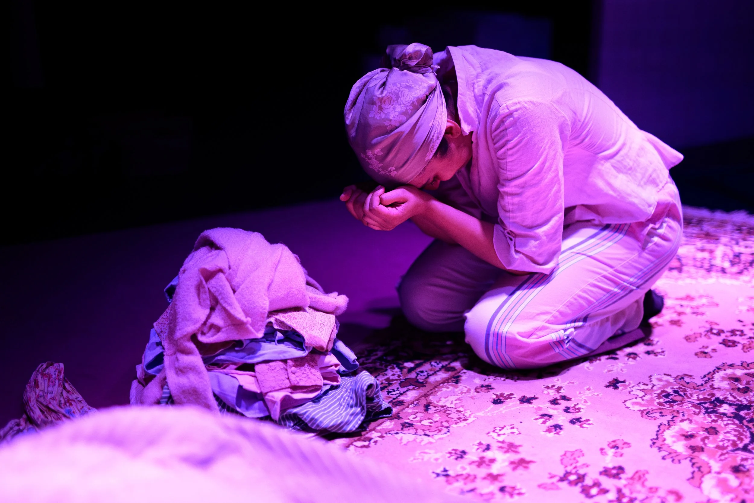 Person wearing a headscarf kneeling on the floor, holding their head in their hands in a room with purple lighting, with clothes and a patterned carpet around them.
