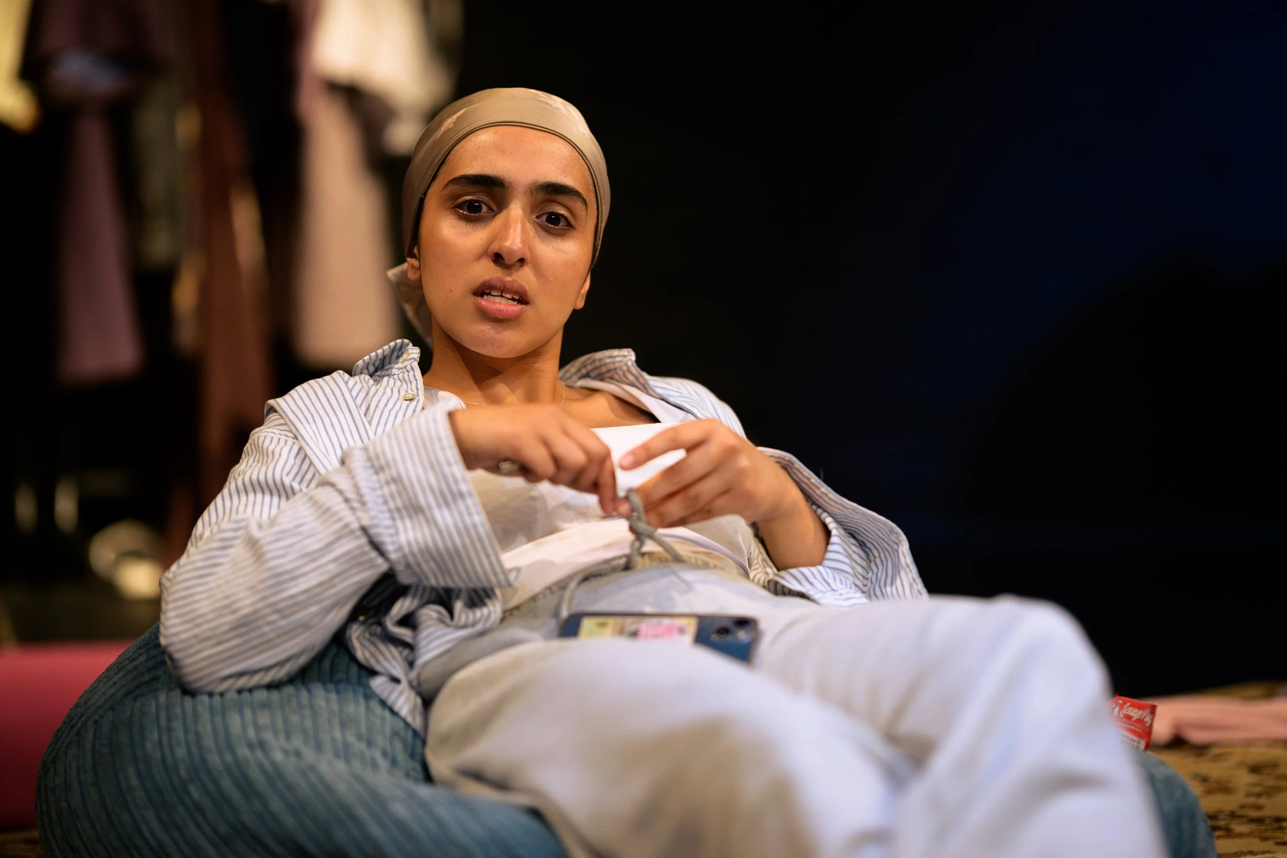 Woman sitting on a bed, wearing a beige headscarf and striped shirt, holding a white bag, with a dark background and blurred closet or wardrobe in the background.