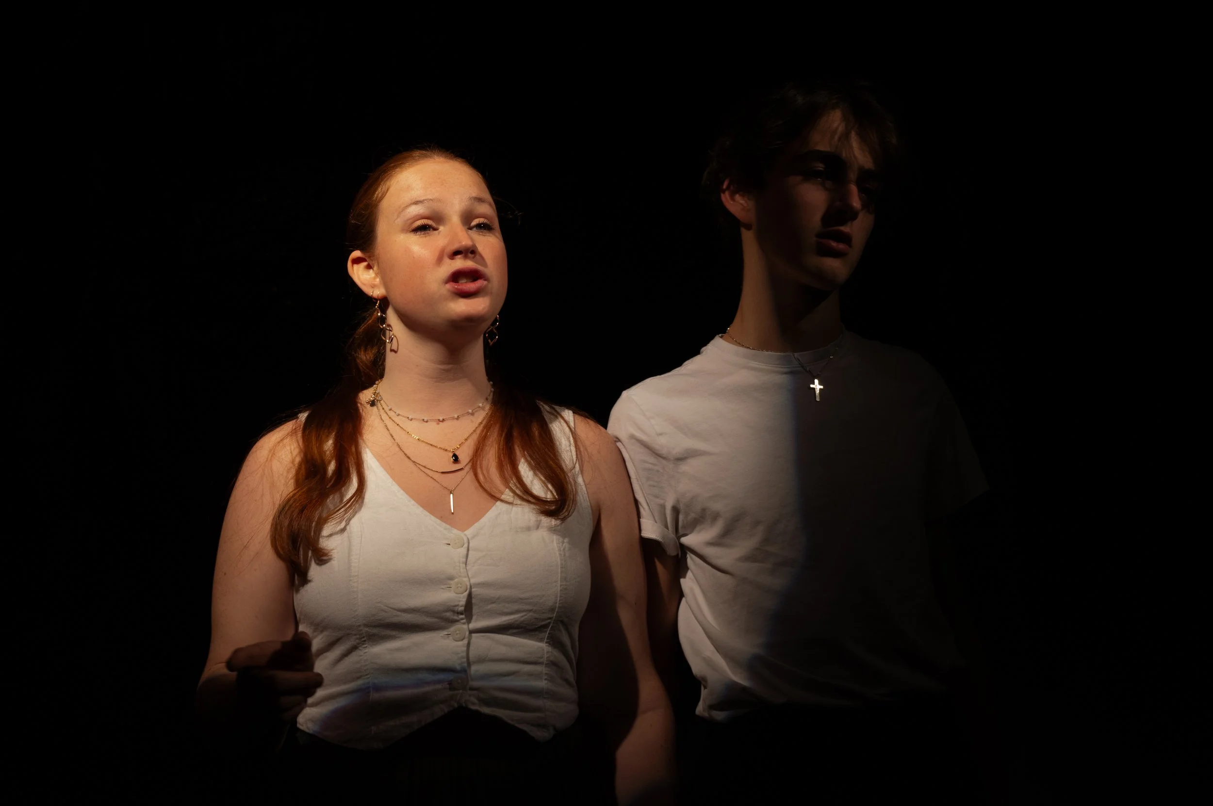Two young people, a woman with red hair and a man with dark hair, standing side by side against a black background, with the woman slightly in front of the man. The woman is wearing a white sleeveless top, layered necklaces, and earrings, and appears