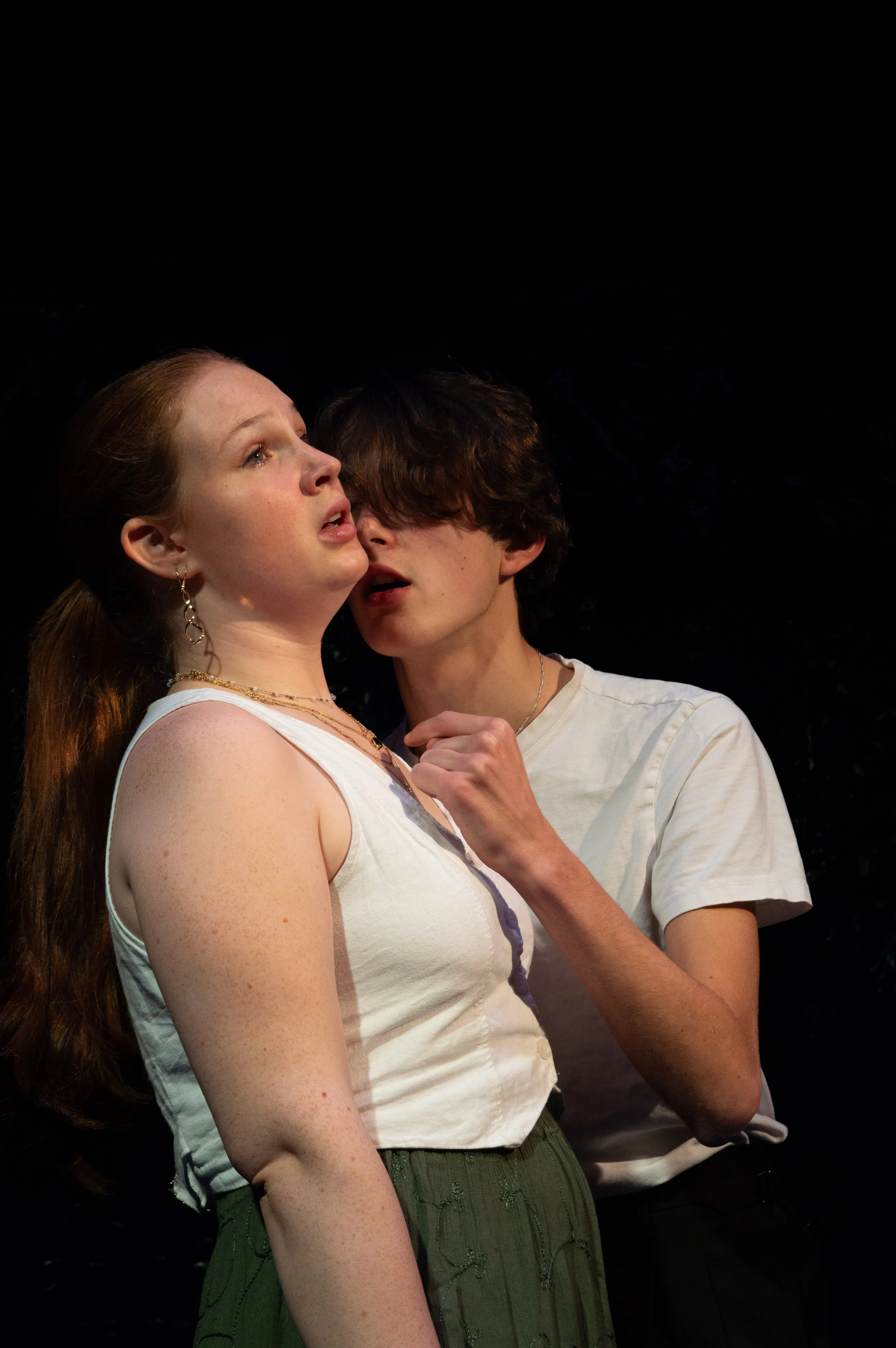 A young woman with red hair and freckles and a young man with dark hair are close together, with the woman's face turned upward and the man leaning in toward her, both appearing to be singing or speaking passionately.