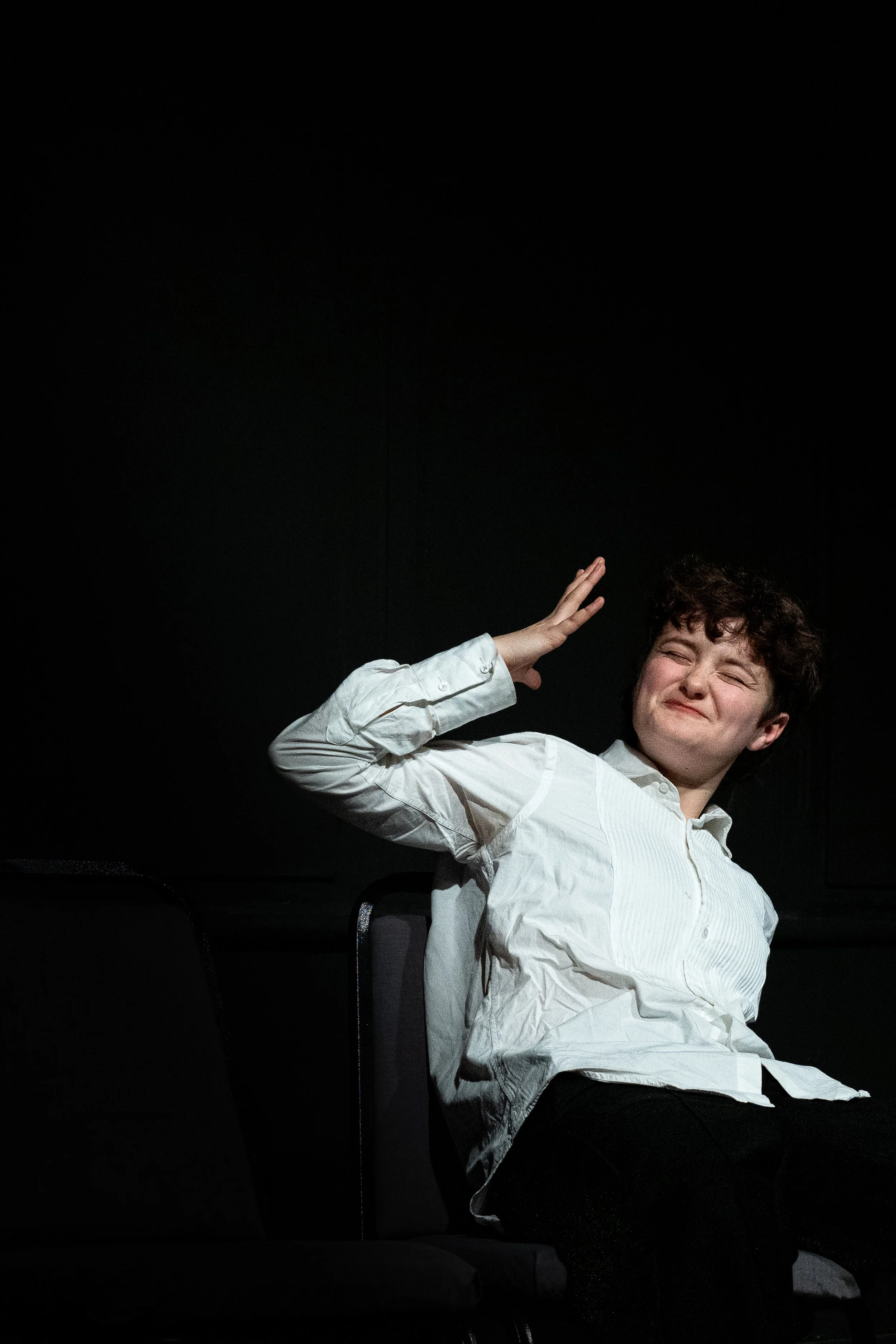 A woman with short curly hair smiling and sitting in a chair, wearing a white button-up shirt, in front of a black background. She is raising her right hand to her forehead in a playful gesture.