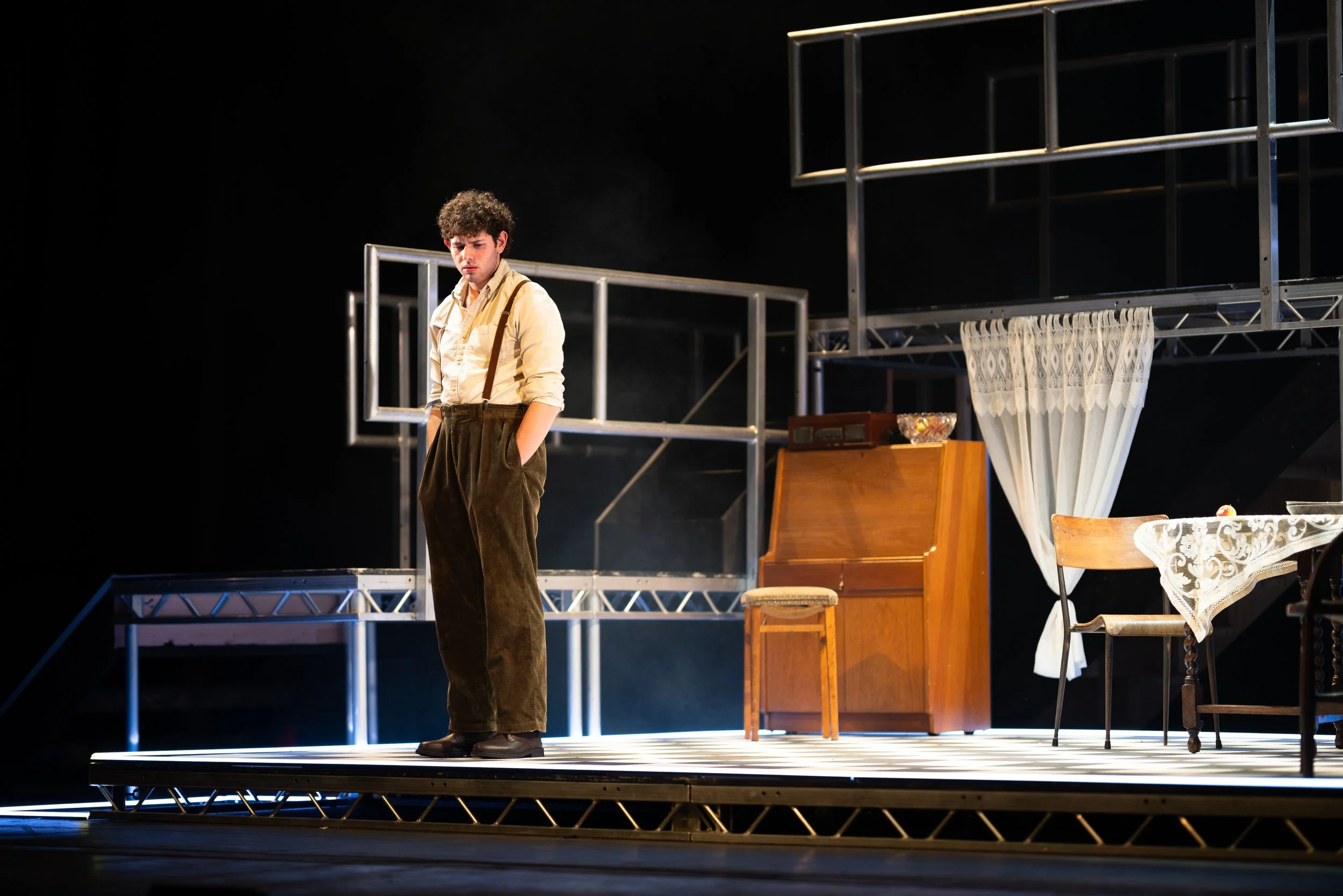 A young man standing on a theatrical stage, looking down thoughtfully, with minimal lighting and a set that includes a table with a lace tablecloth, a chair, a wooden cabinet, and metal framework in the background.