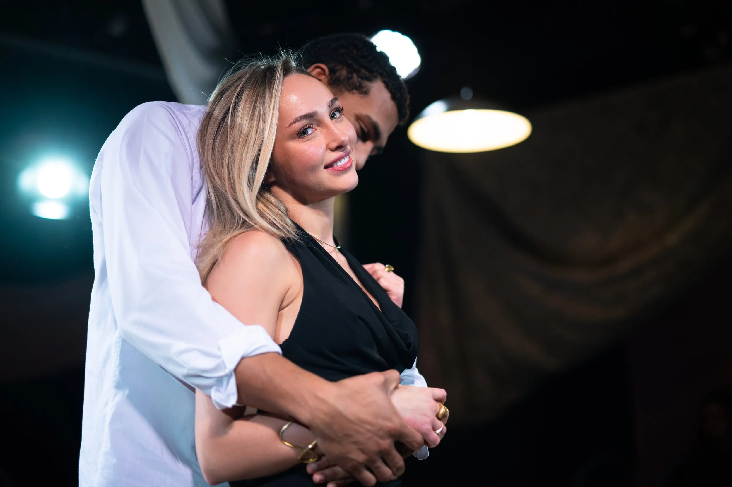 A man in a white shirt embracing a woman in a black sleeveless top, both with hands on her stomach, in a dark setting with ceiling lights.