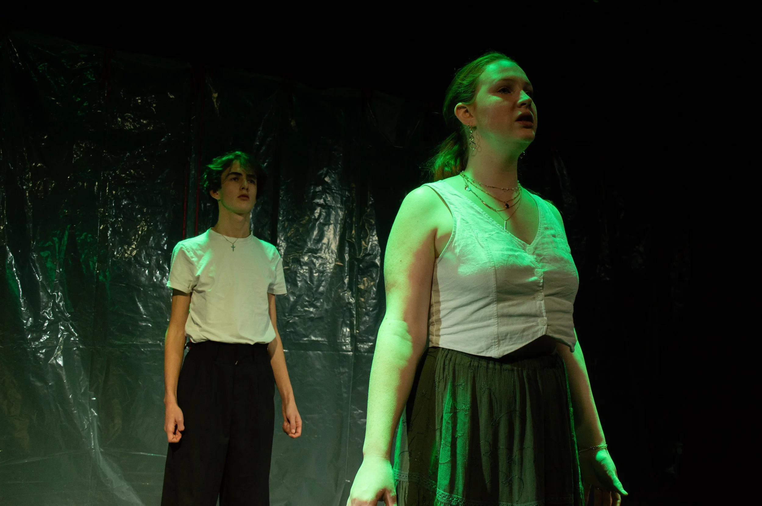 Two young women stand on a stage with dark background and green lighting, wearing white tops and dark bottoms, facing opposite directions.