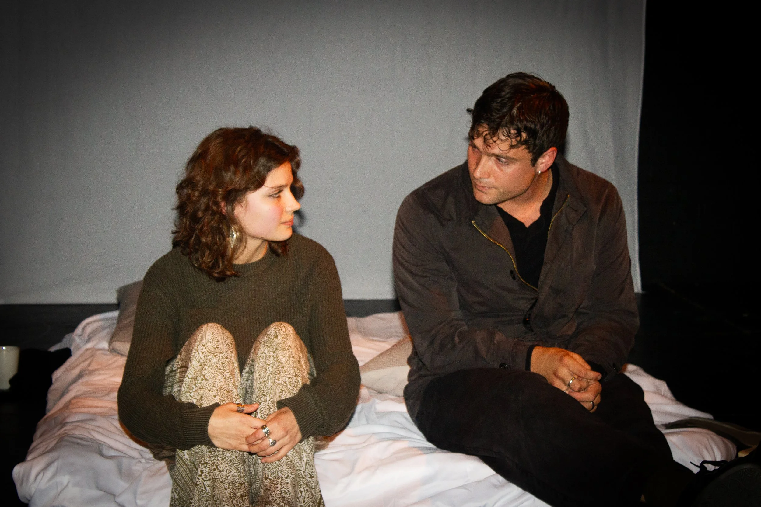 A young woman with wavy dark hair and a young man with dark wavy hair sitting on a bed, looking at each other with serious expressions, in a dimly lit room.