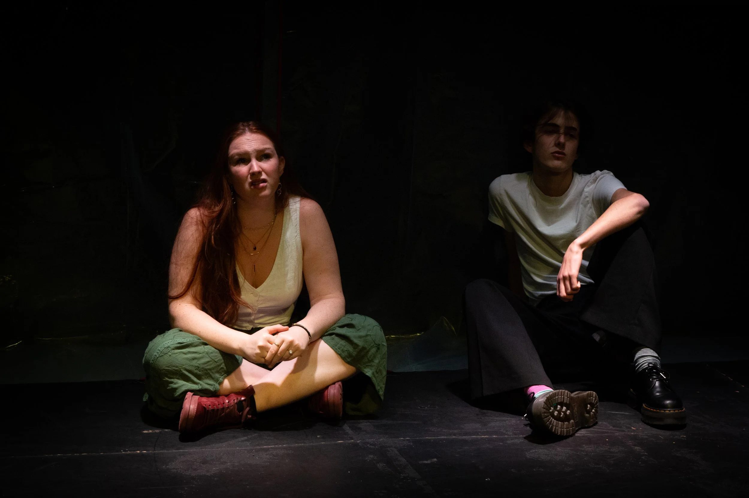Two young actors sitting on a dark stage, engaged in a performance or rehearsal, illuminated by stage lighting.