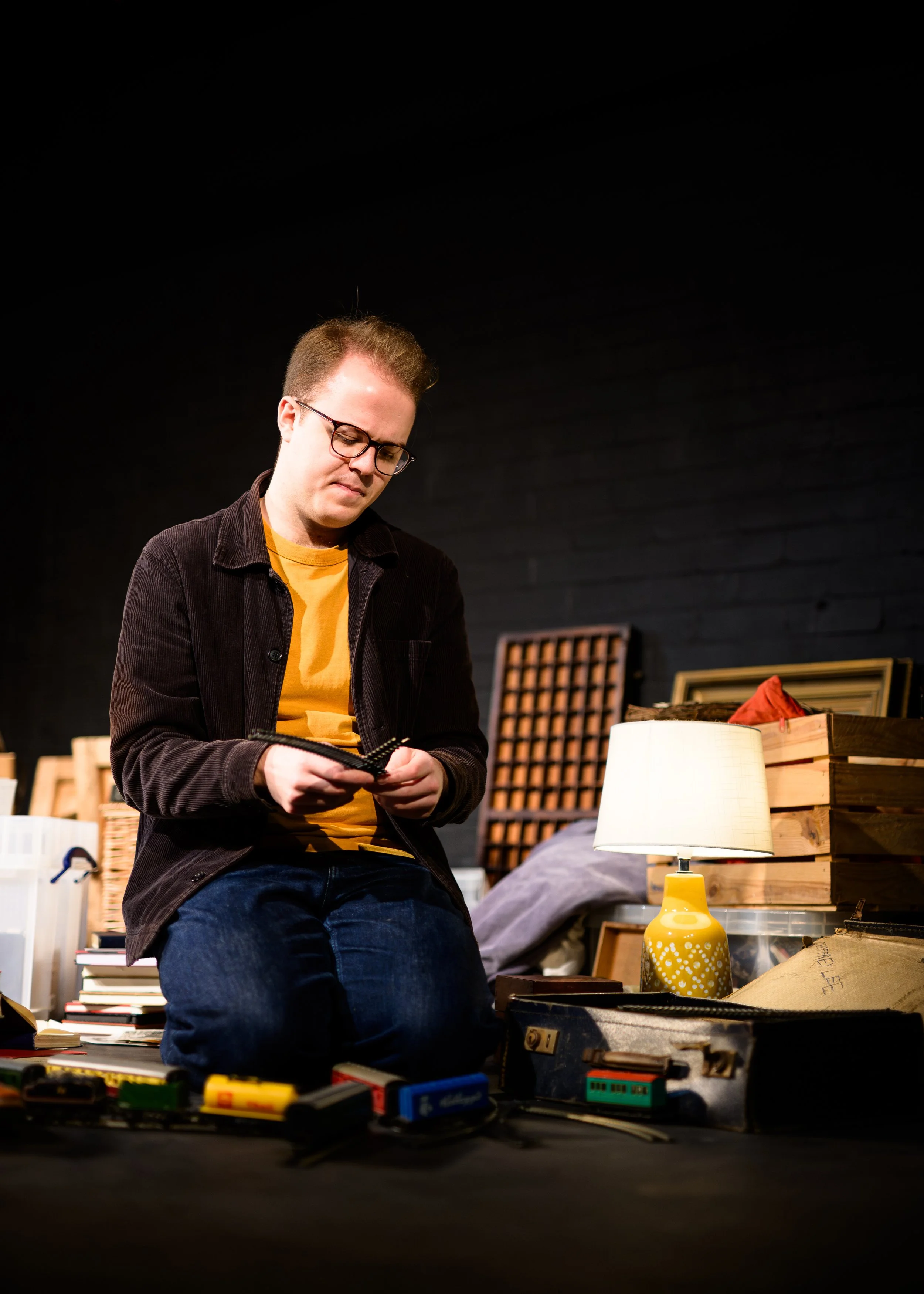 A young man with glasses, wearing a yellow shirt and dark jacket, sitting on a bench in a room filled with books, boxes, and a yellow lamp.