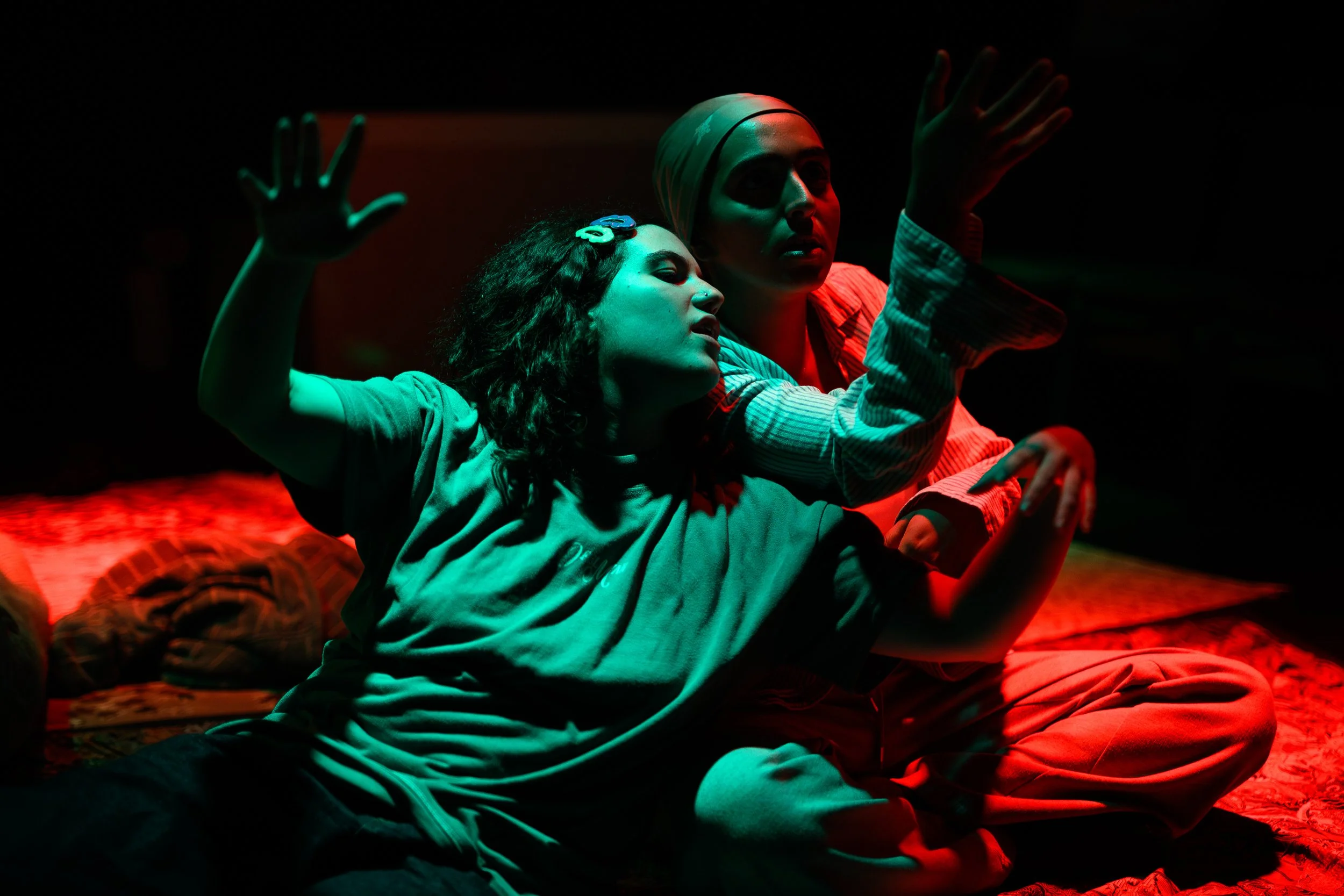 Two young women sitting close together under dark lighting with red and green hues. The woman in the foreground has curly hair, a hair clip, and a nose piercing, and is gesturing with her hands. The woman behind her is wearing a headscarf and striped