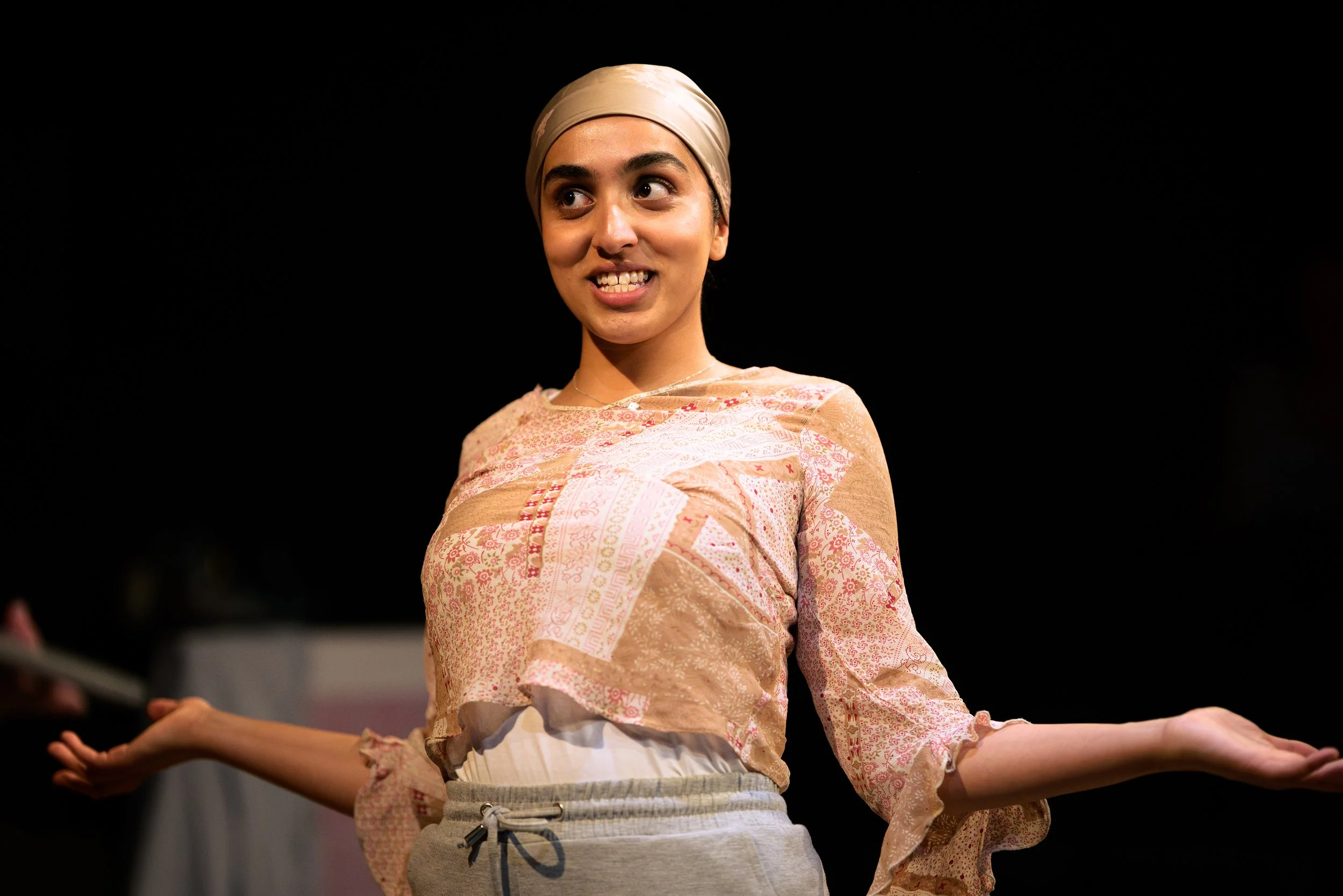 A woman wearing a beige headscarf and a pink patterned blouse stands with arms outstretched against a dark background with stage lighting.