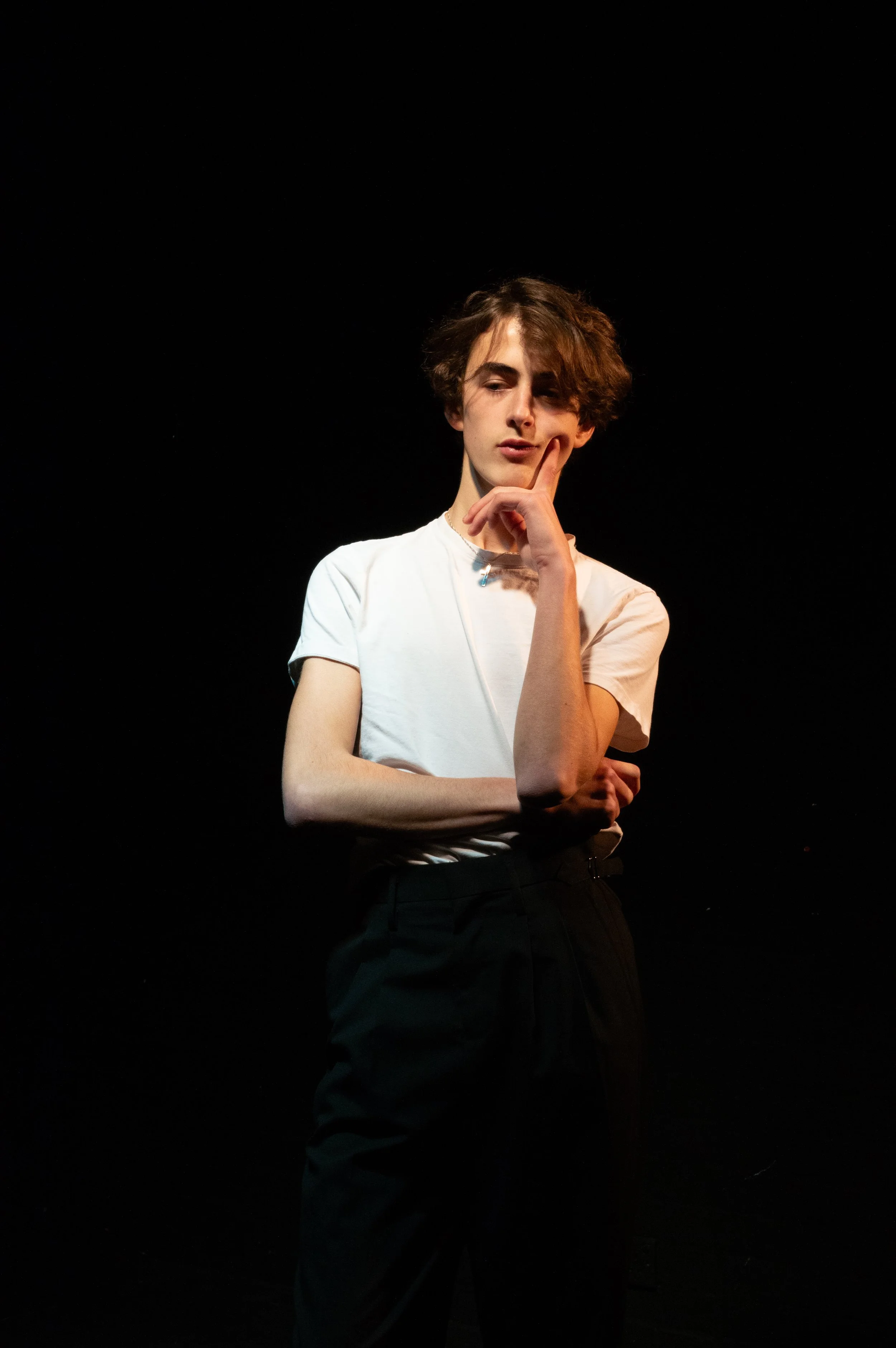 A person with short, wavy brown hair, wearing a white t-shirt and dark pants, standing against a solid black background, with one hand touching their face in a thoughtful pose.